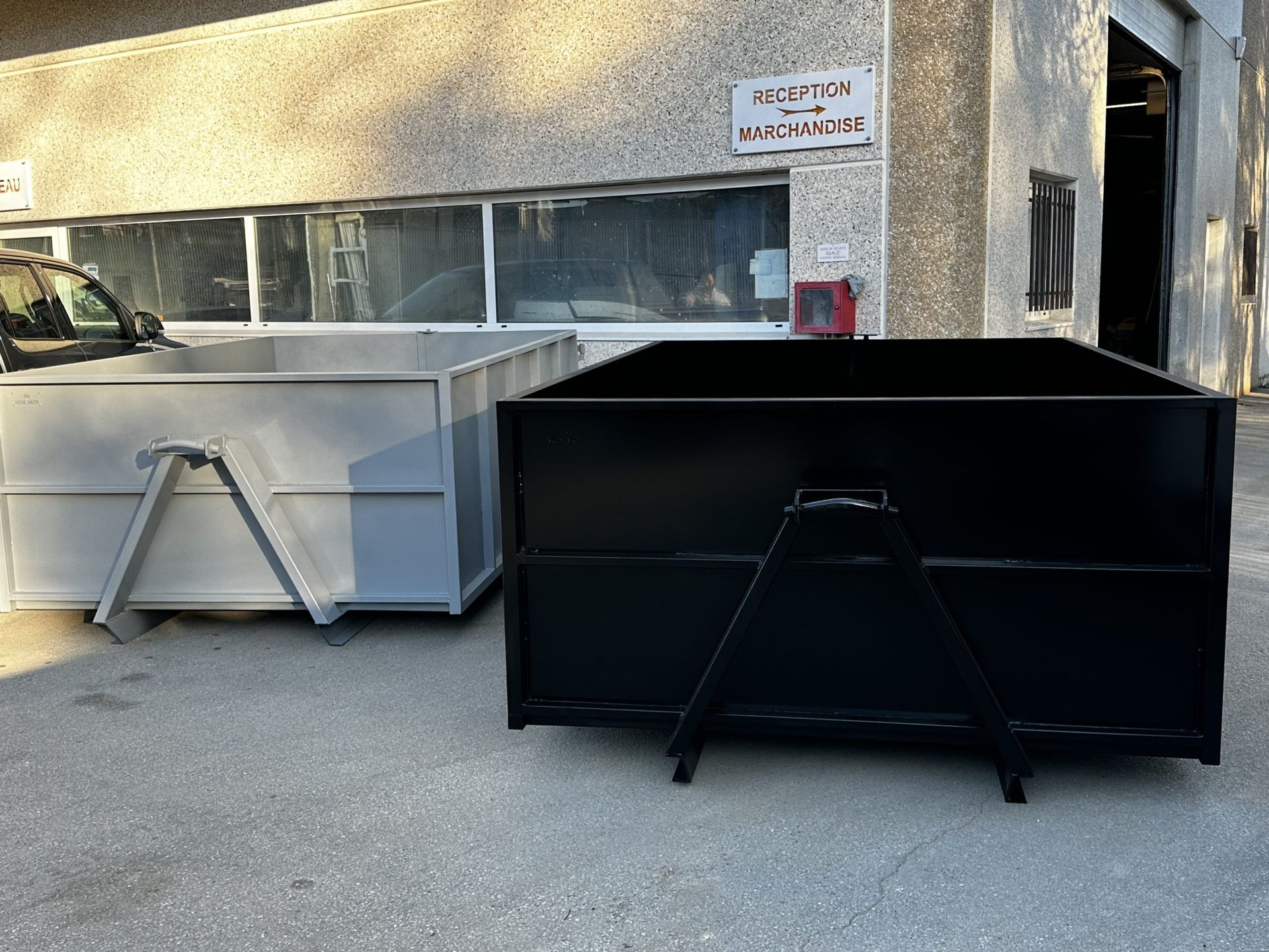Deux grandes poubelles métalliques à roulettes, l'une argentée et l'autre noire, à l'extérieur d'un bâtiment, sous une fenêtre.