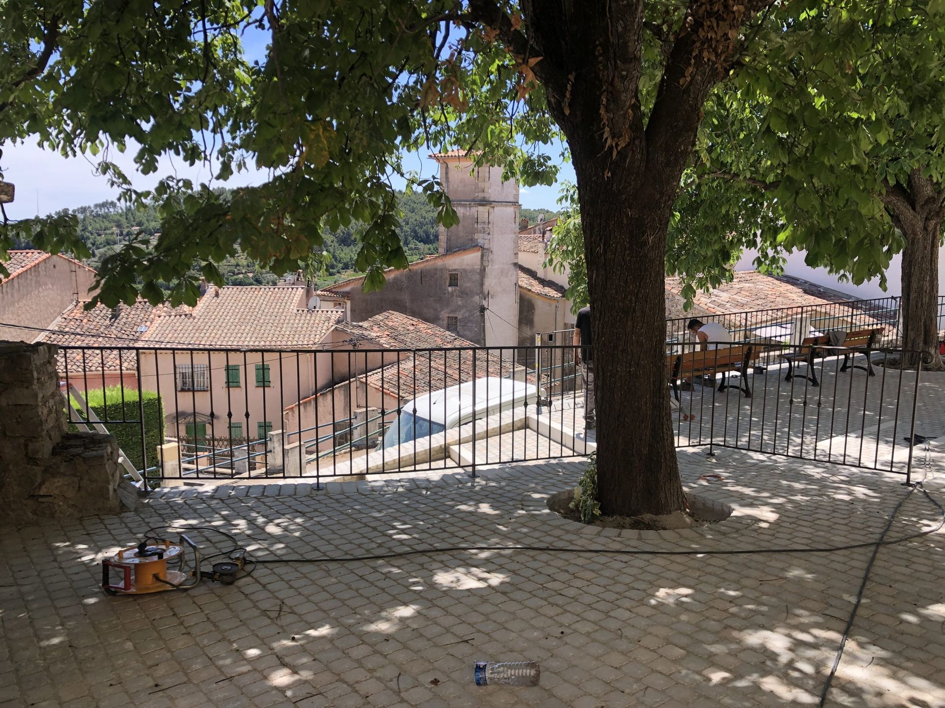 Cour intérieure ombragée avec arbres, pavage en pierre, clôture en fer forgé et toits de tuiles rouges en arrière-plan