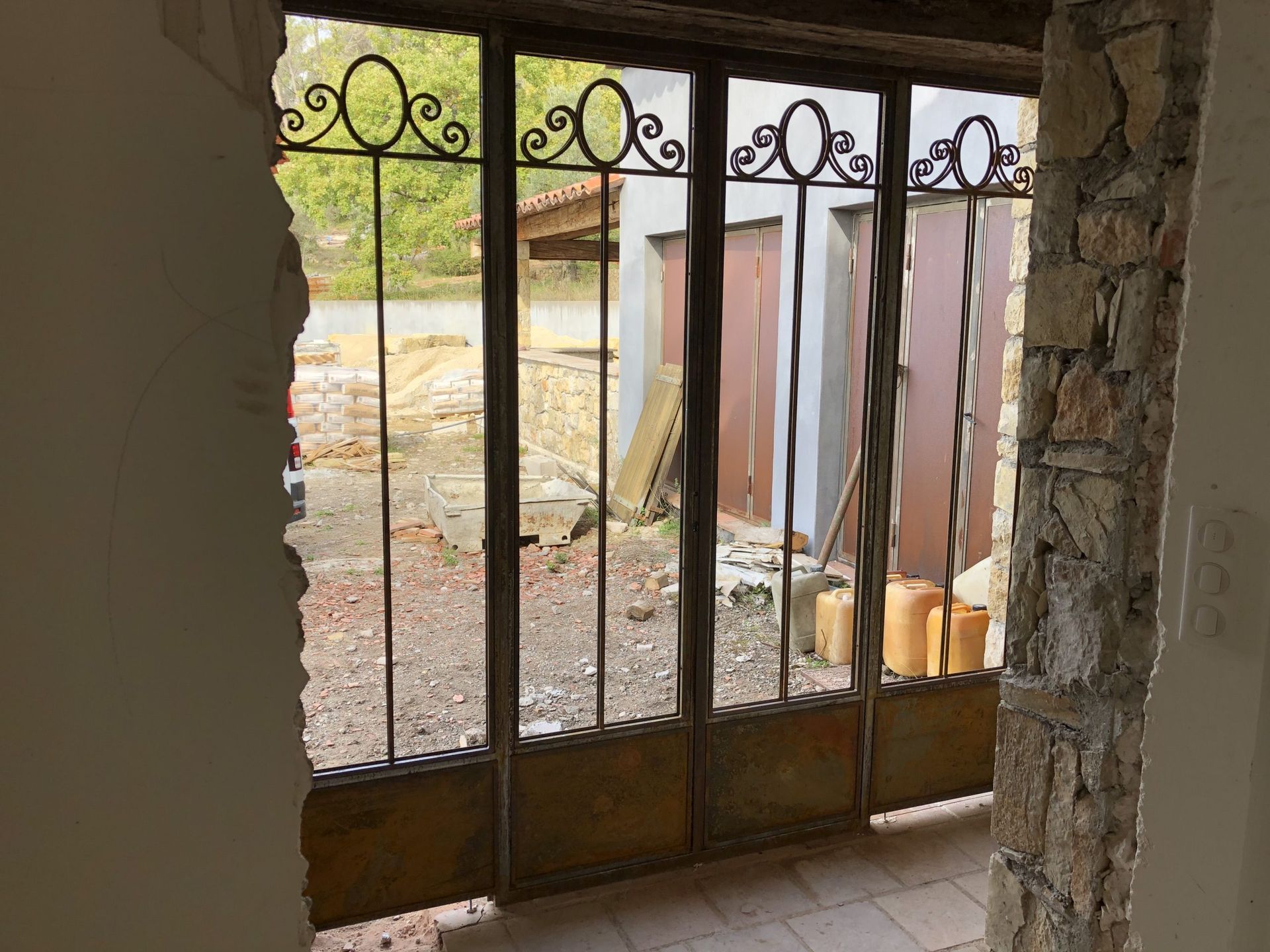 Un portail en fer forgé, à l'intérieur d'une porte en pierre, s'ouvre sur une cour de gravier ensoleillée avec des tissus suspendus à l'extérieur.