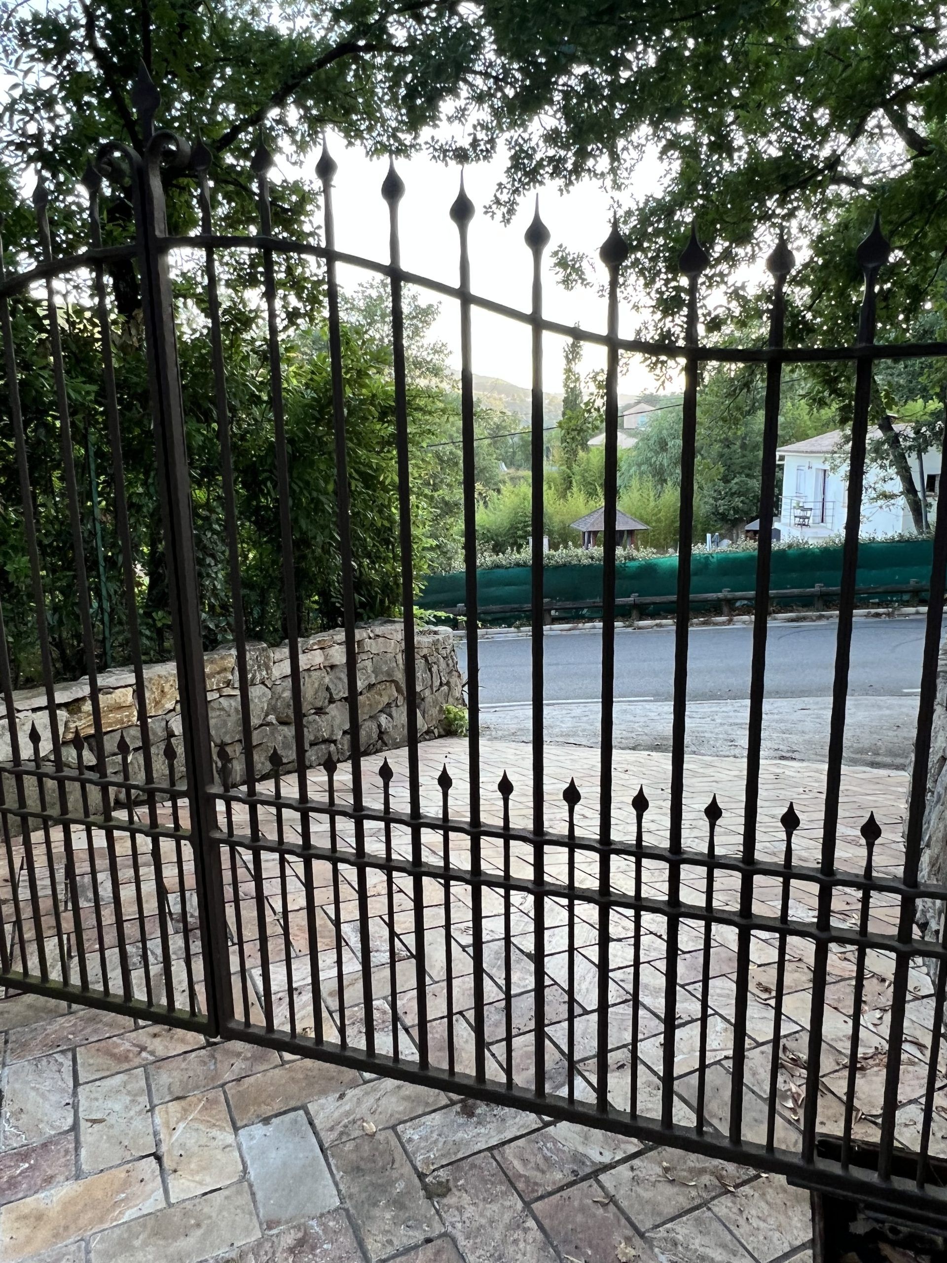 Un portail en fer noir devant une allée pavée, avec des arbres et une rue au-delà.