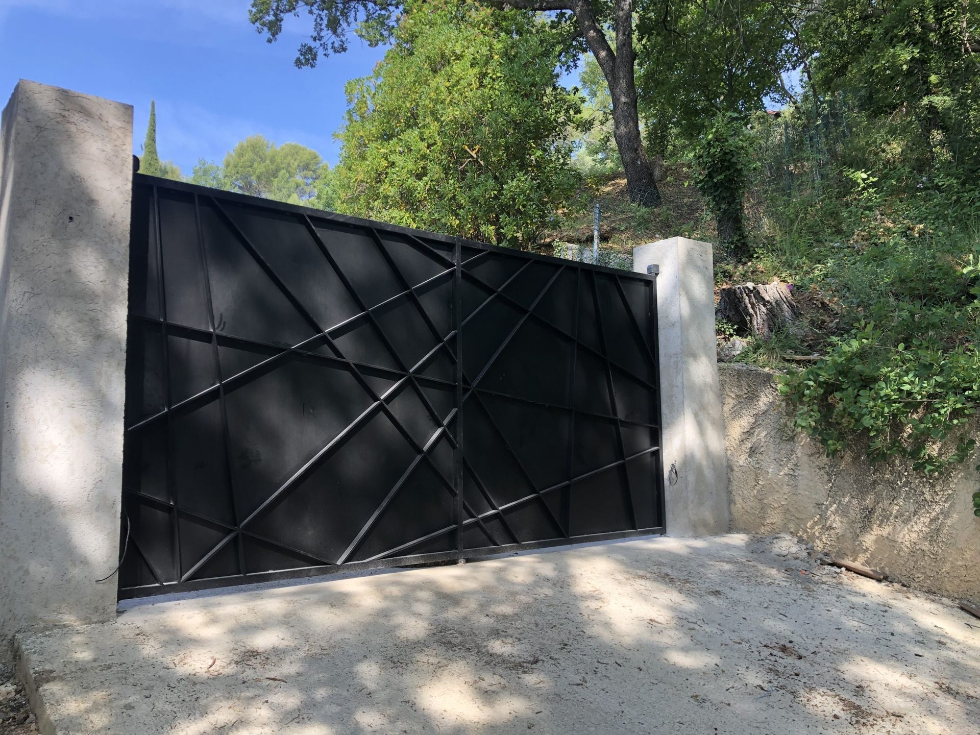 Portail en métal noir entre des piliers en béton, sur une allée de terre ensoleillée, avec des arbres et une colline en arrière-plan.