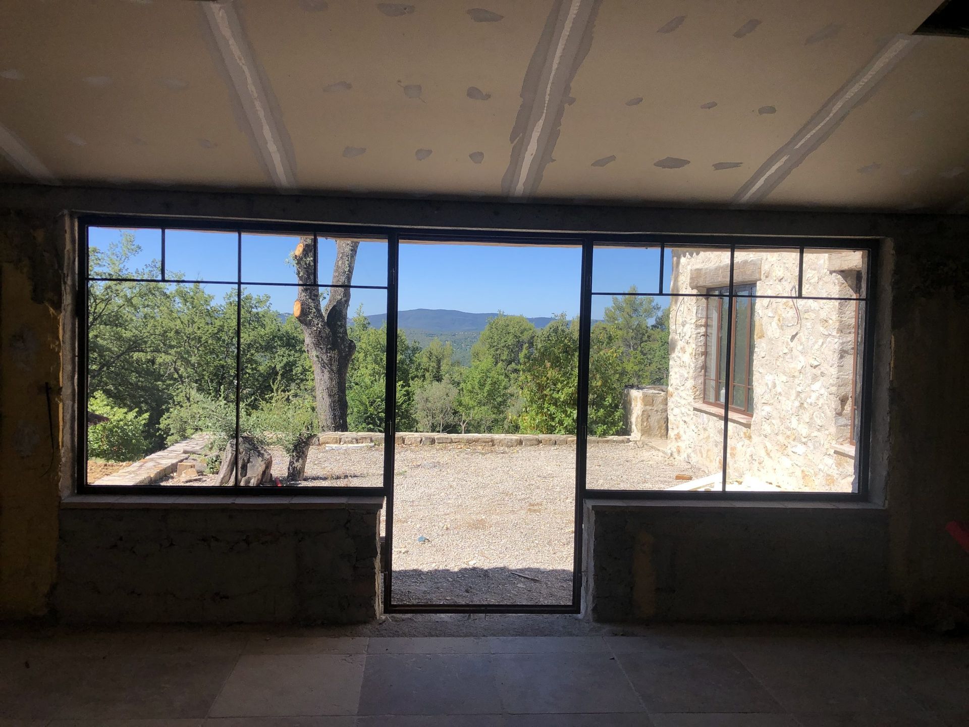 Pièce intérieure vide avec une large fenêtre et une porte donnant sur une cour extérieure ensoleillée et arborée.