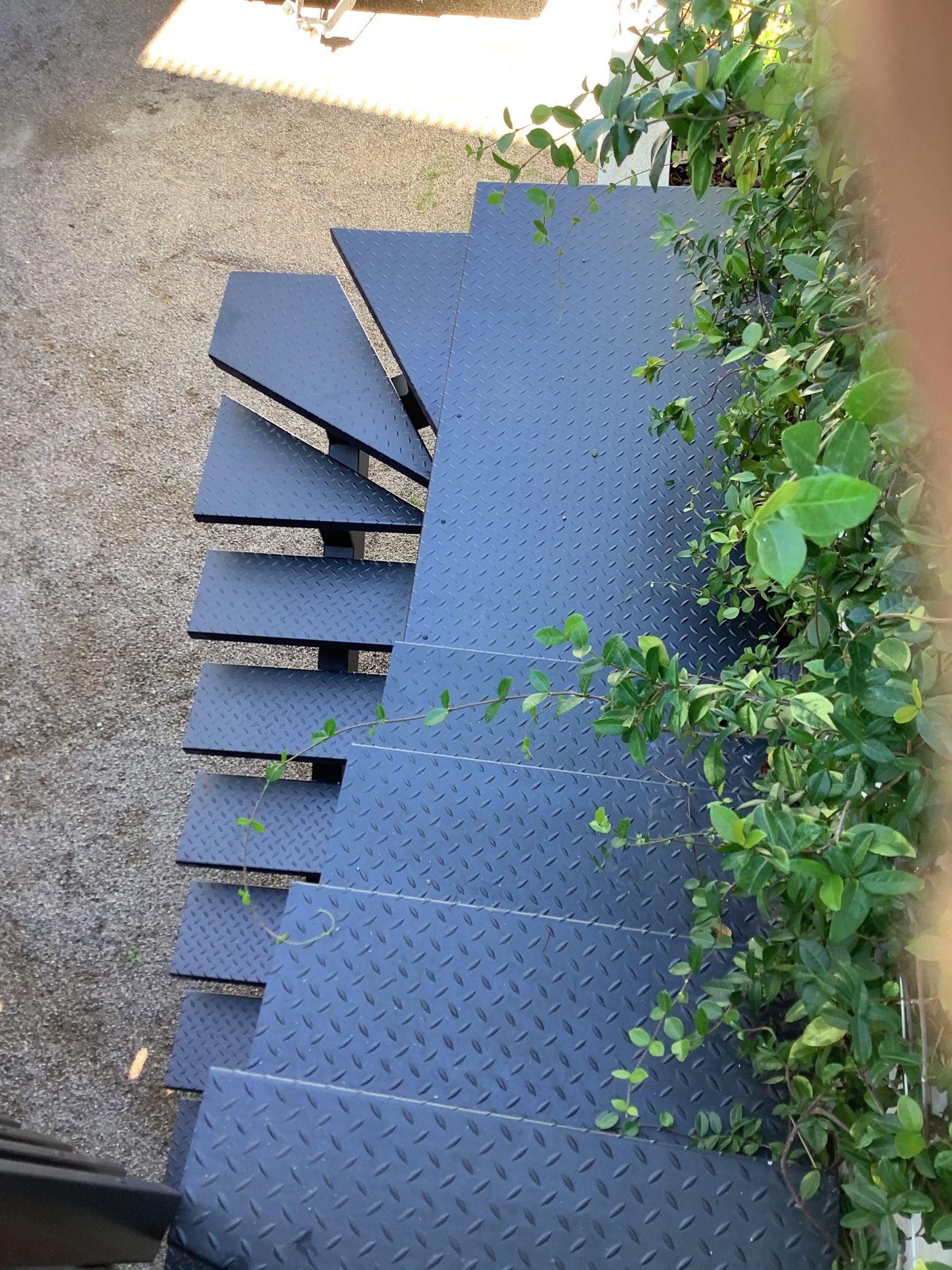 Escalier en colimaçon métallique bleu à côté d'une haie verte, vu d'en haut.