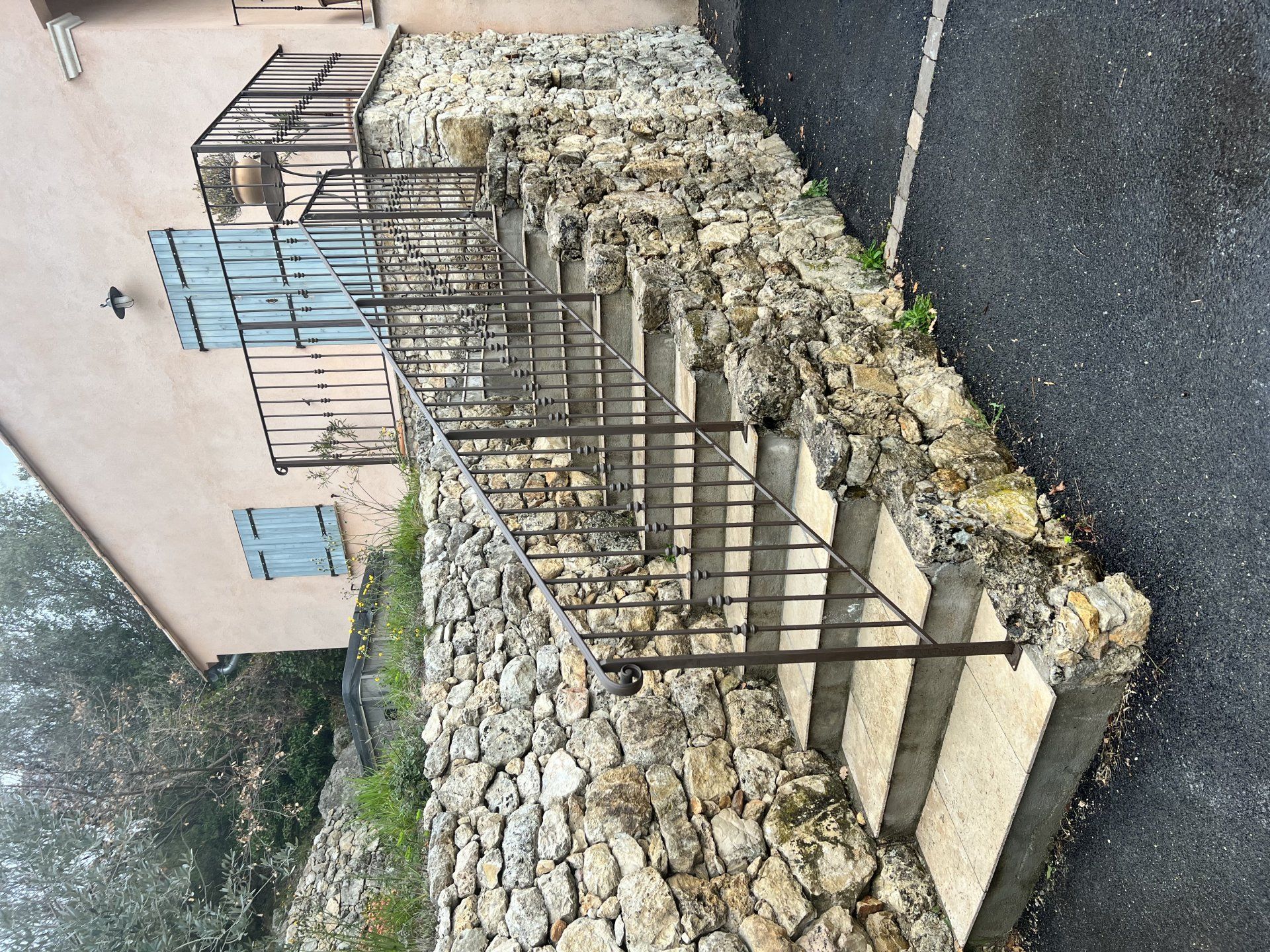 Mur en pierre avec rambarde métallique jouxtant un bâtiment beige et une rue sombre, vu sous un certain angle