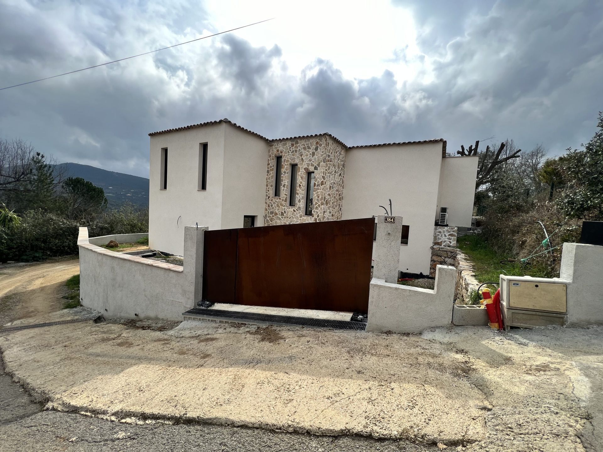 Maison blanche moderne avec un portail marron et un muret, vue depuis une allée de gravier sous un ciel nuageux