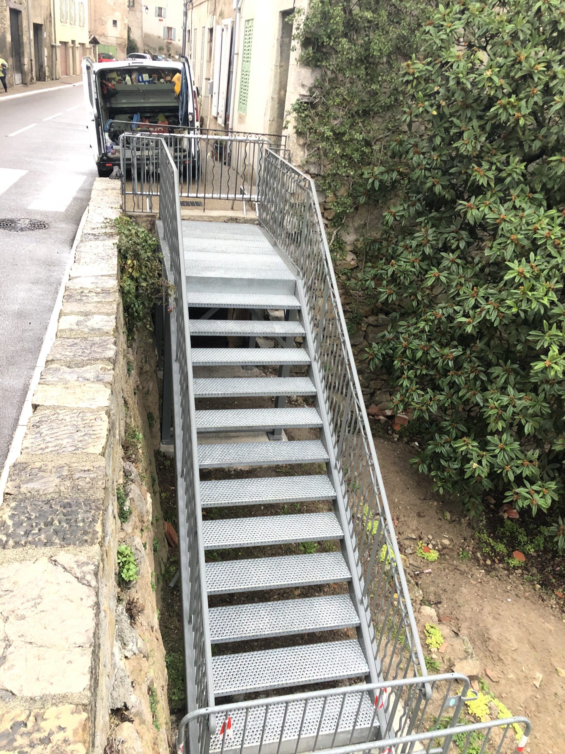 Un escalier extérieur abrupt, entre des murs de pierre et de la verdure, avec un véhicule noir garé en haut.