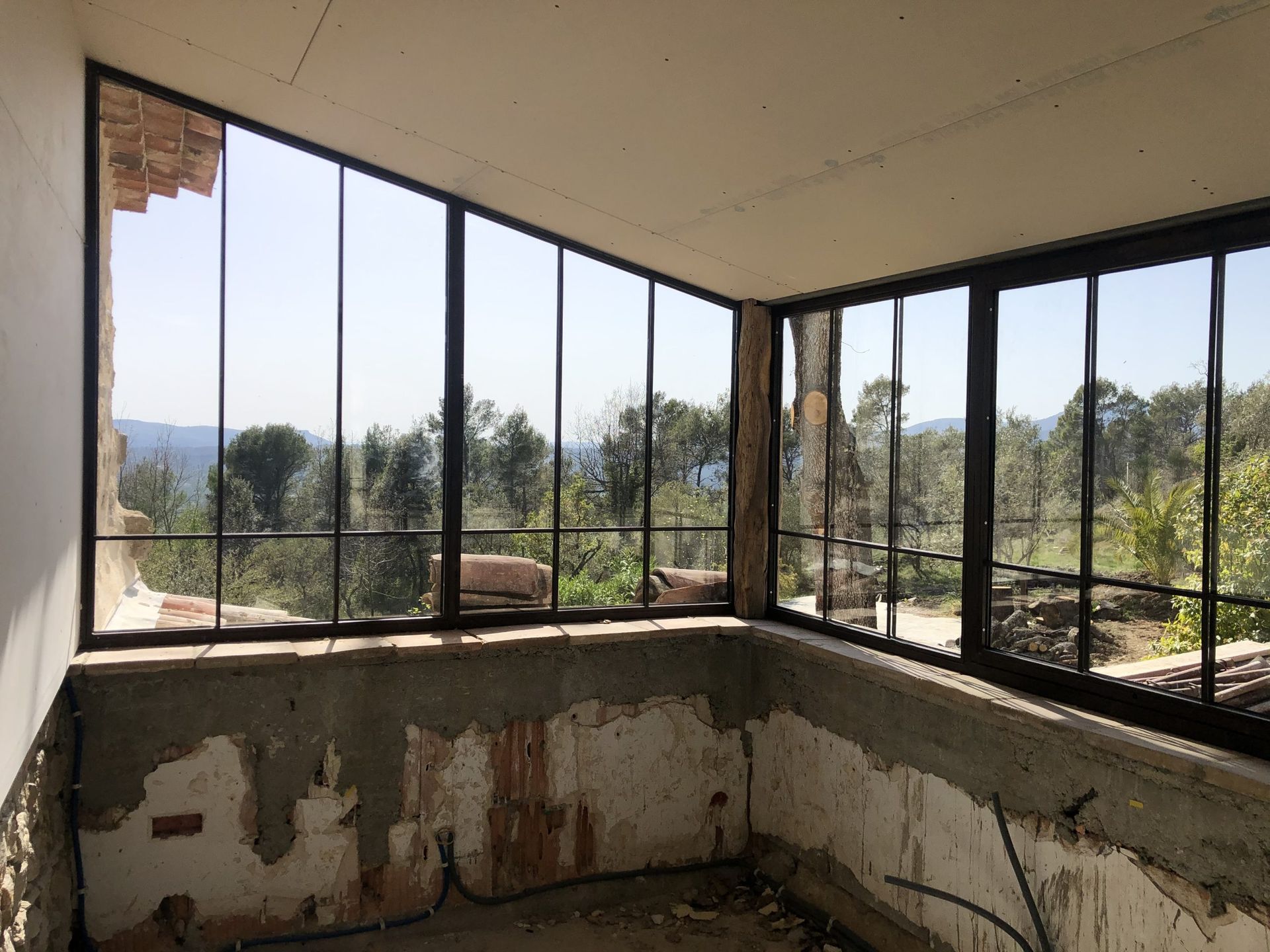 Pièce abandonnée avec des fenêtres brisées, des murs qui s'écaillent et une vue sur les arbres à l'extérieur.