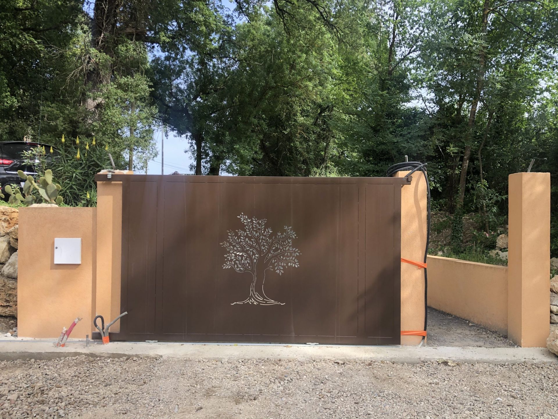 Un portail en métal brun et un mur en stuc beige avec une petite pancarte, situés devant des arbres feuillus sous la lumière vive du jour.