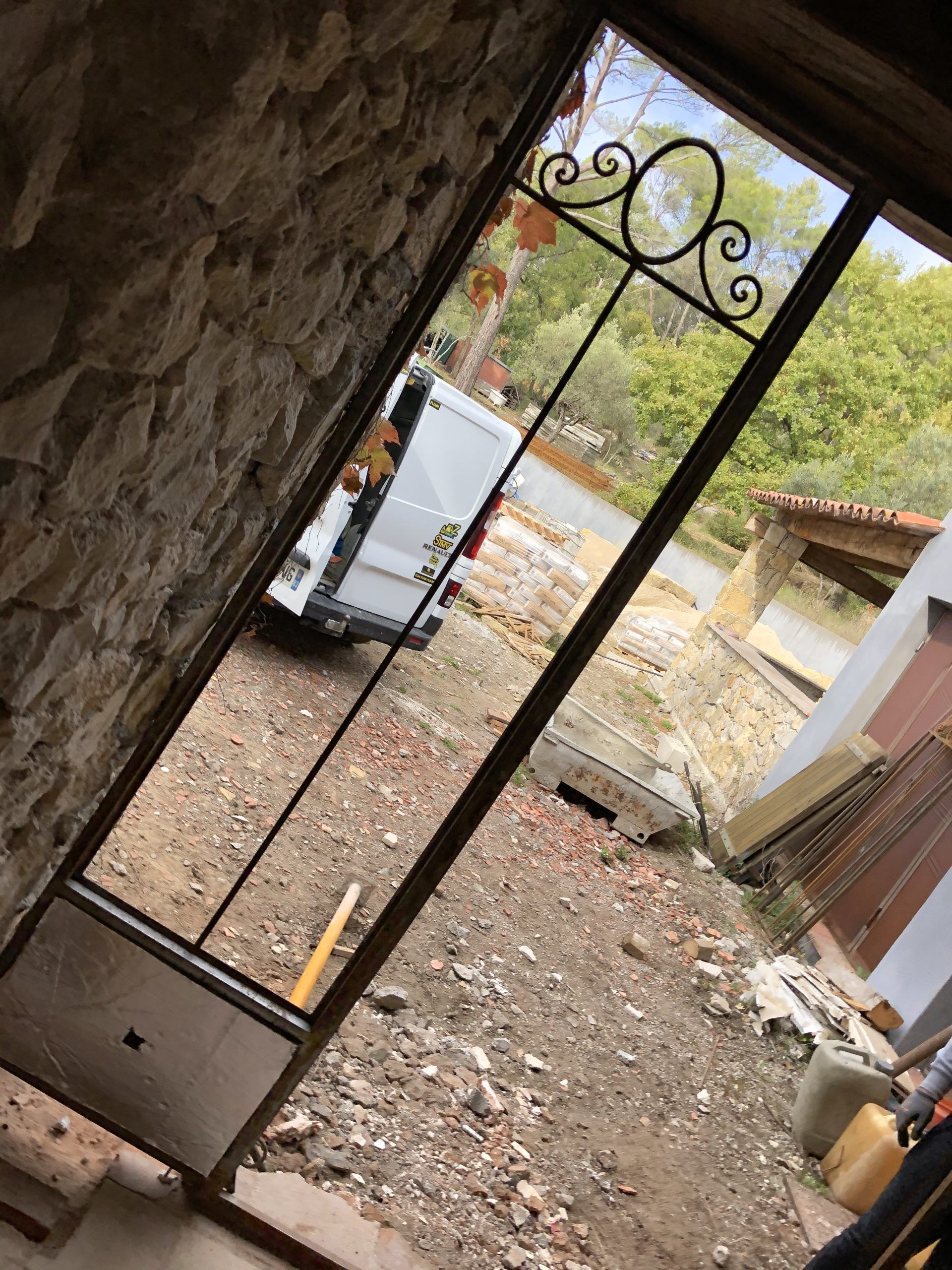 Vue en plongée à travers un portail métallique donnant sur une cour gravillonnée avec une camionnette blanche et un abri de jardin à l'extérieur.