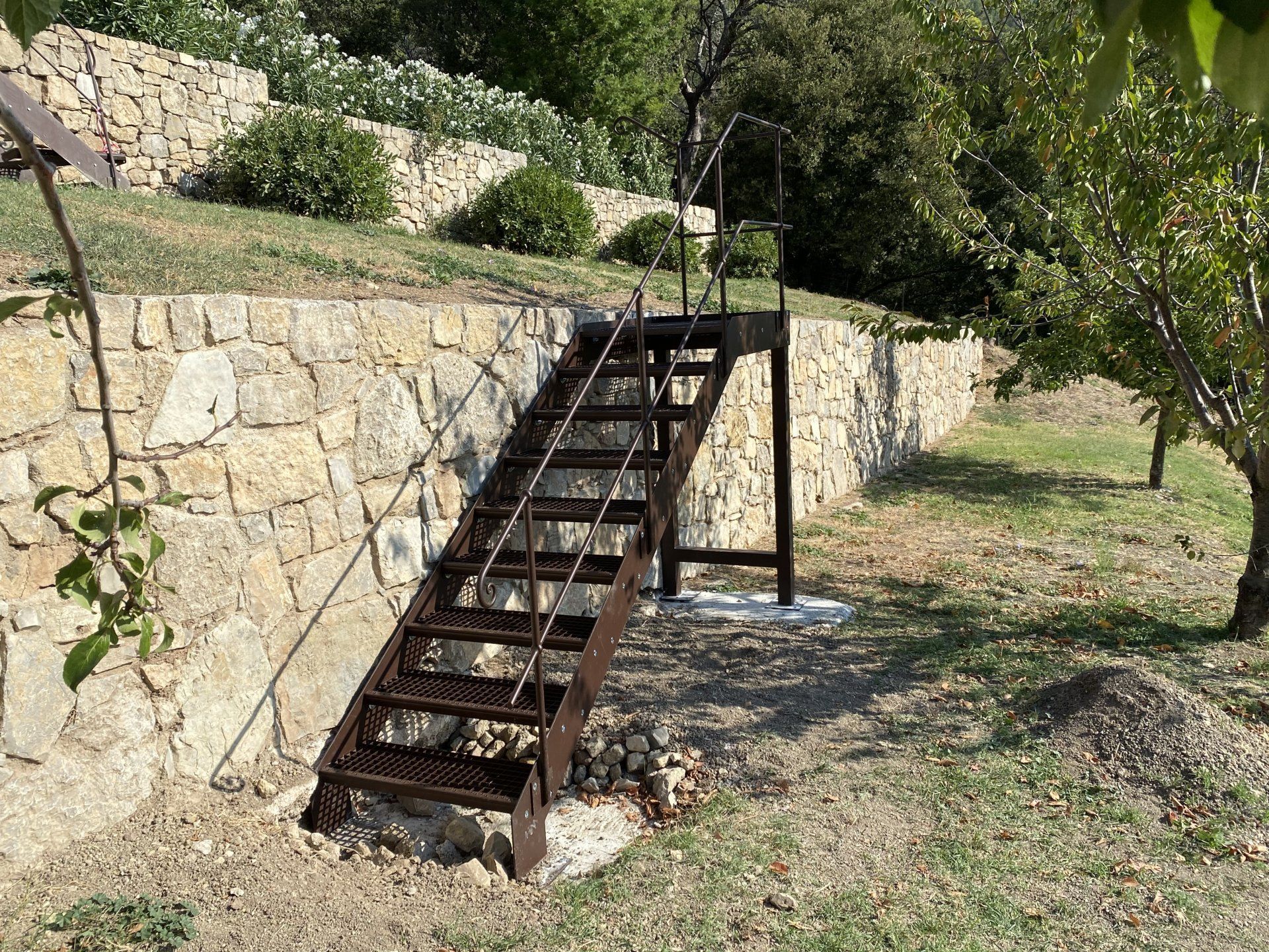Escalier en métal rouillé à côté d'un mur de pierre dans un espace extérieur herbeux et arboré.