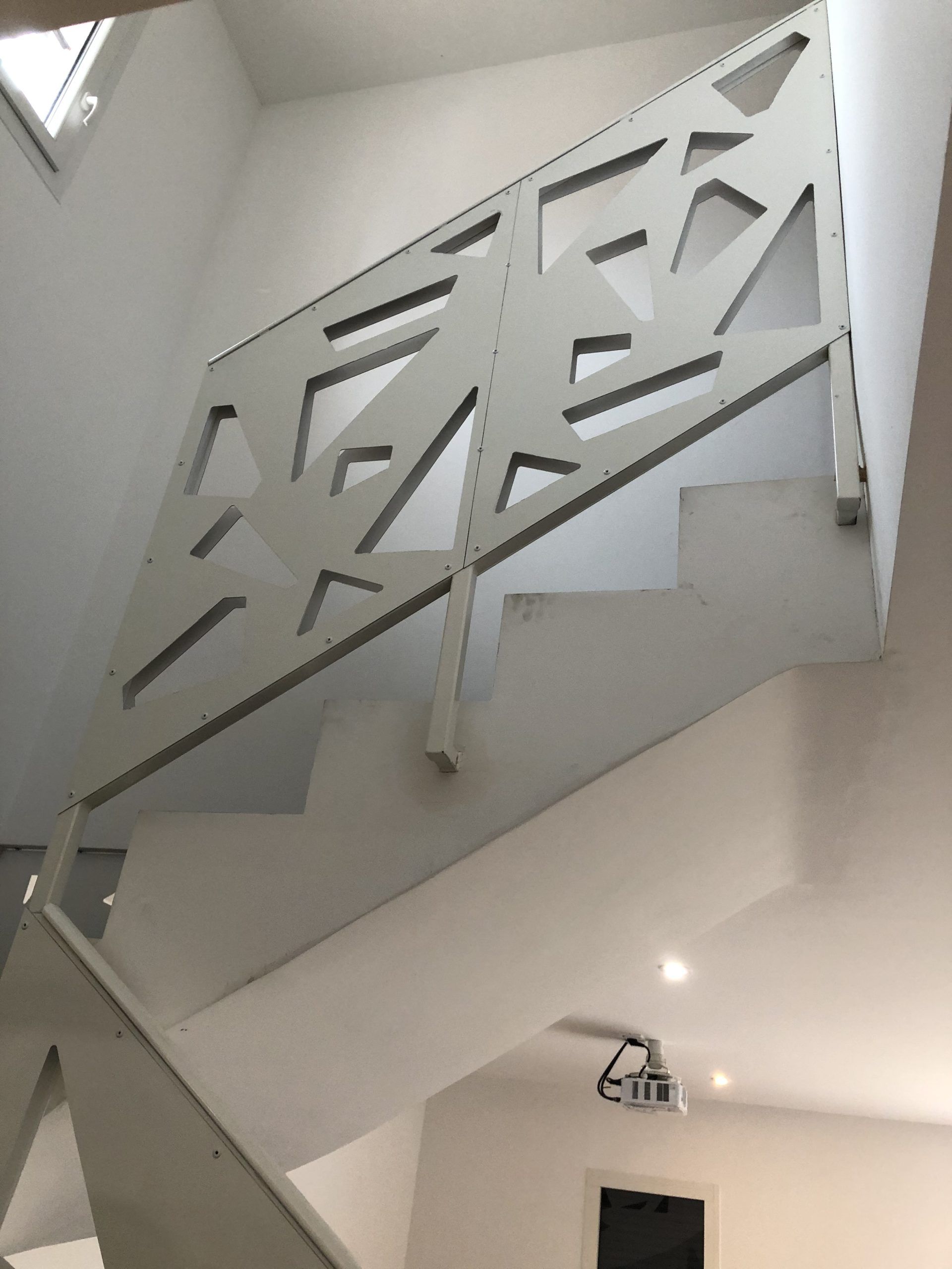 Escalier intérieur avec une rampe en métal gris ornée de découpes géométriques, vu d'en bas.
