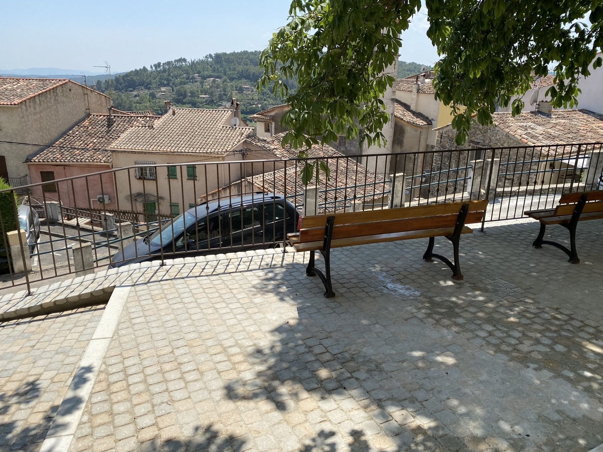Terrasse en pierre avec bancs donnant sur un village à flanc de colline et des toits sous les arbres