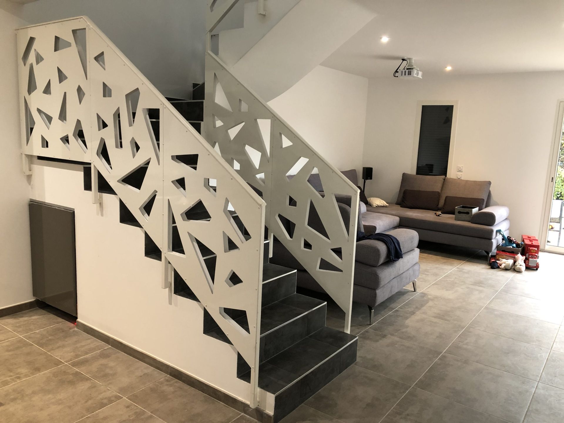 Salon moderne avec un escalier décoratif, un canapé gris et un grand sol carrelé.