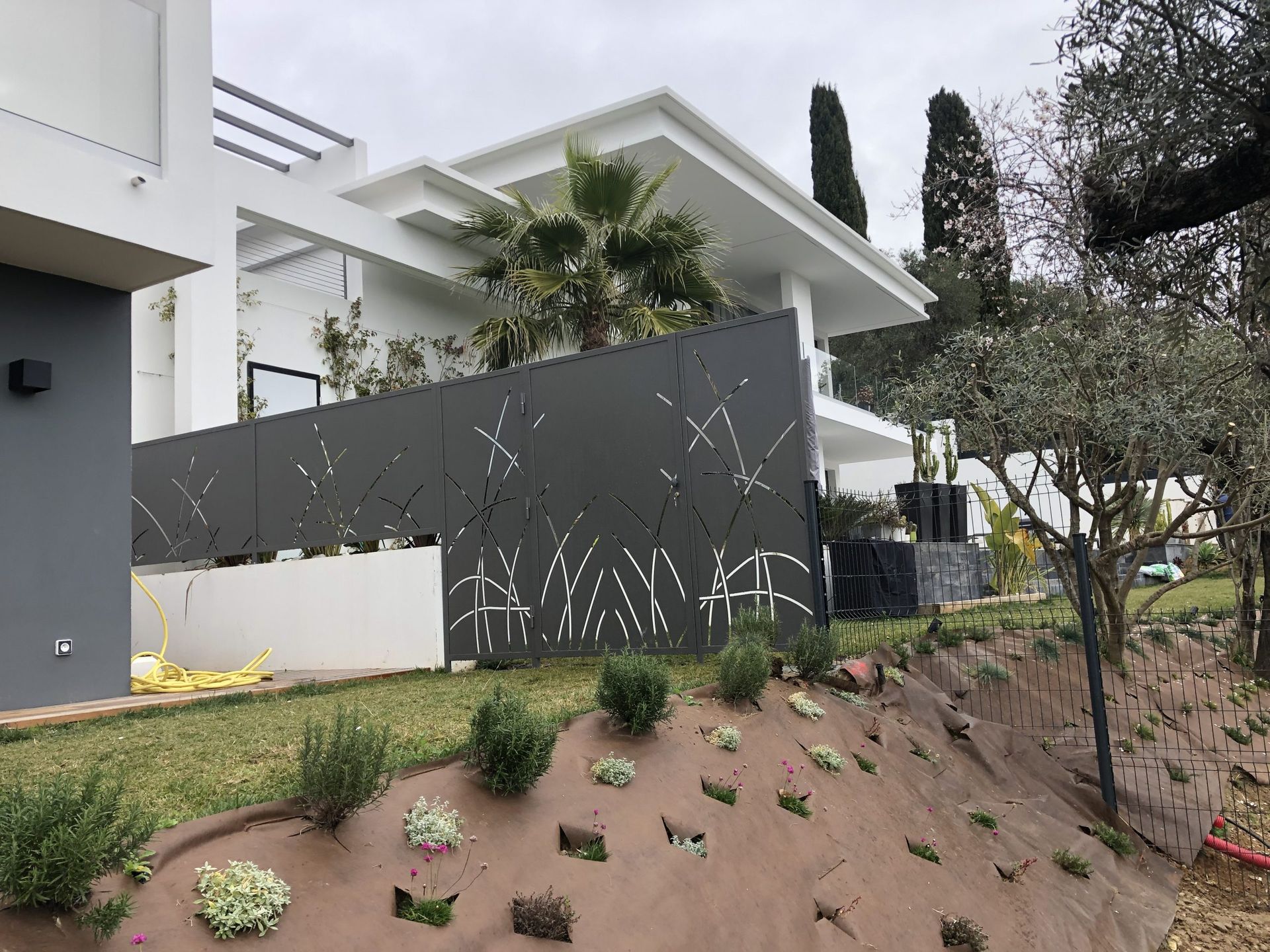 Maison blanche moderne avec une clôture foncée, des arbres et un jardin paysager en pente avec de petites plantes