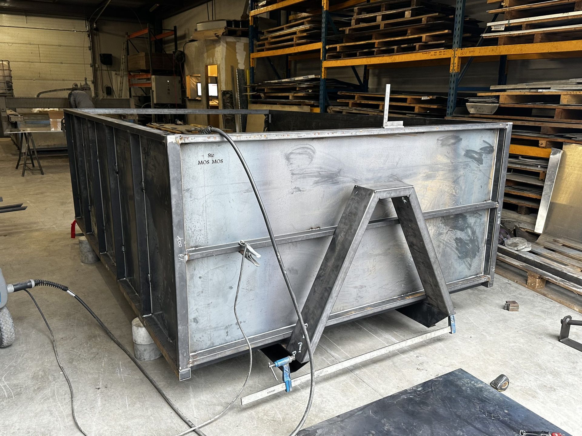 Grand conteneur industriel métallique avec supports de renfort dans un atelier d'entrepôt, entouré de matériaux empilés.
