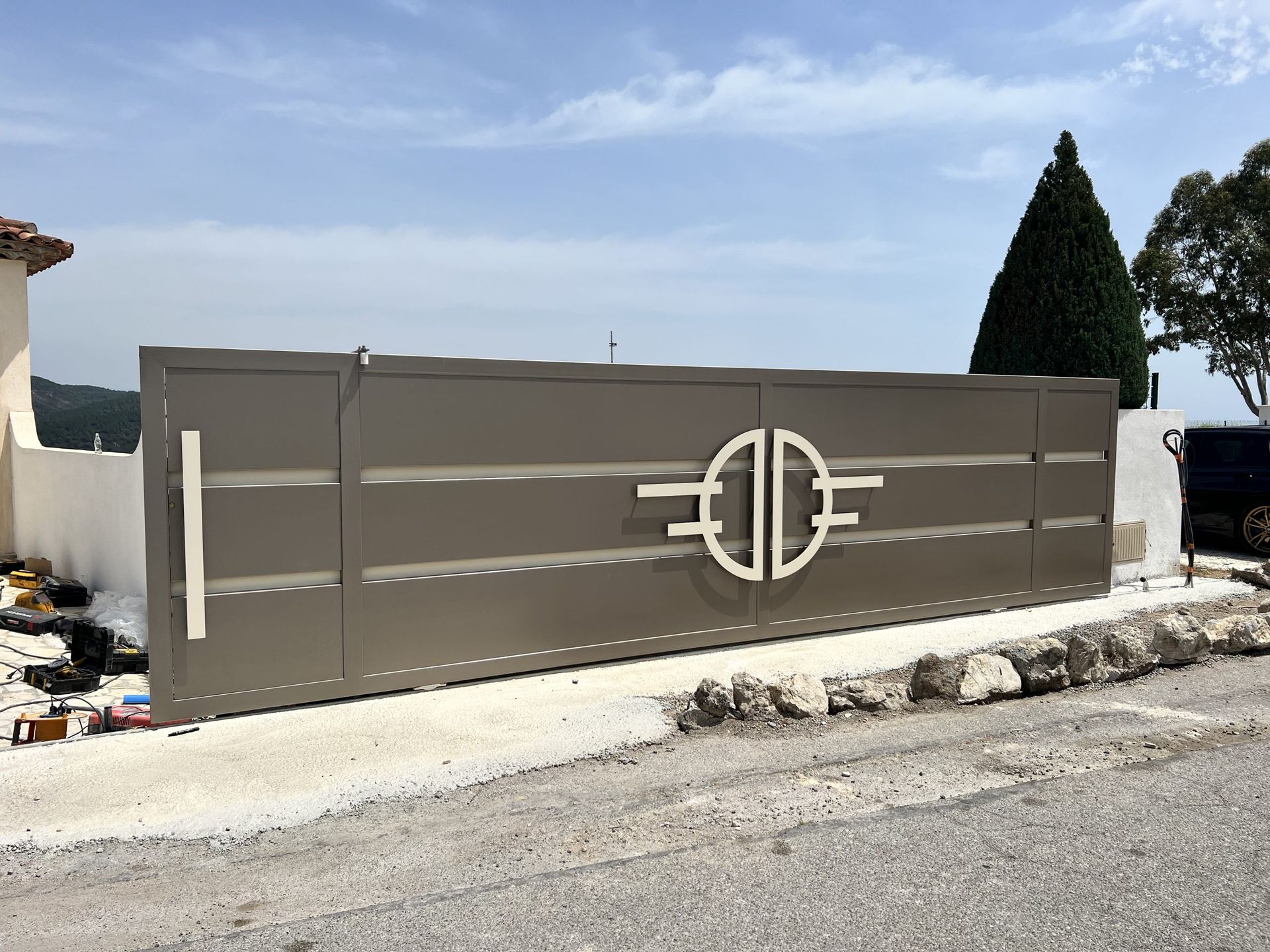 Portail d'allée en métal gris avec un emblème décoratif circulaire, à côté d'un mur en bord de route