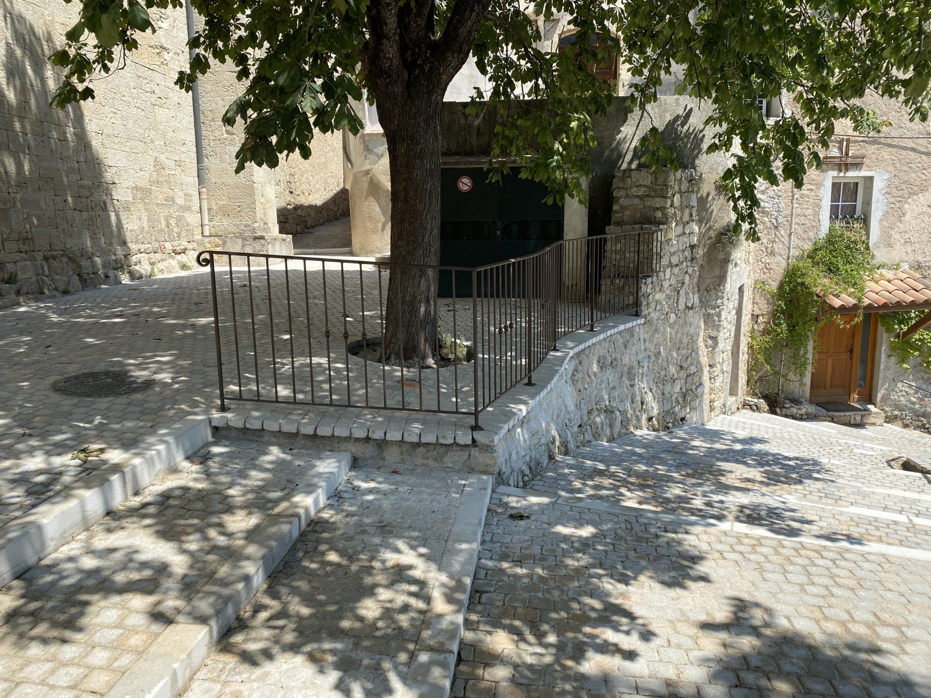 Cour pavée avec un grand arbre, une balustrade en métal et une porte sombre dans la cour ensoleillée d'un vieux bâtiment