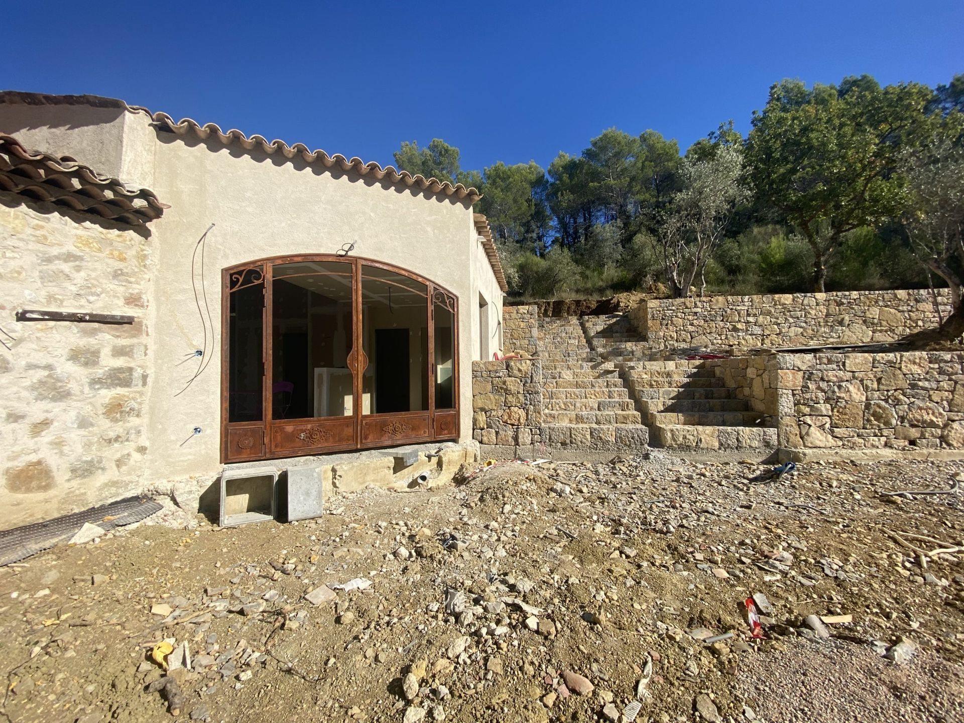 Maison en pierre avec porte cintrée en bois et mur de terrasse sous un ciel bleu clair