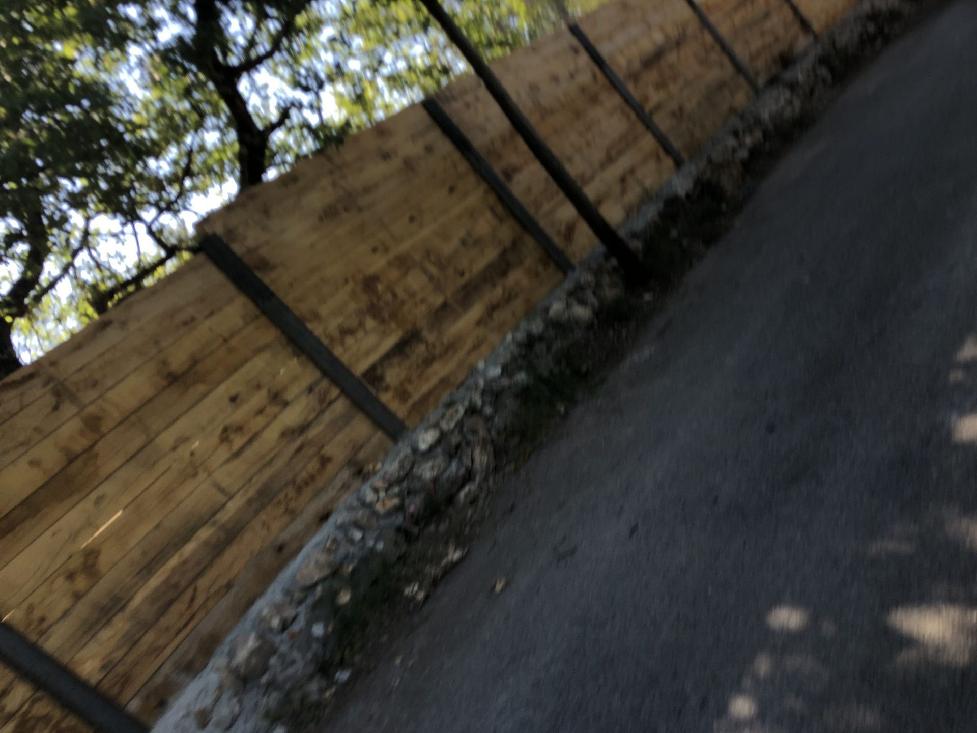 Vue inclinée d'une clôture en bois bordant une route pavée, des arbres projetant des ombres.