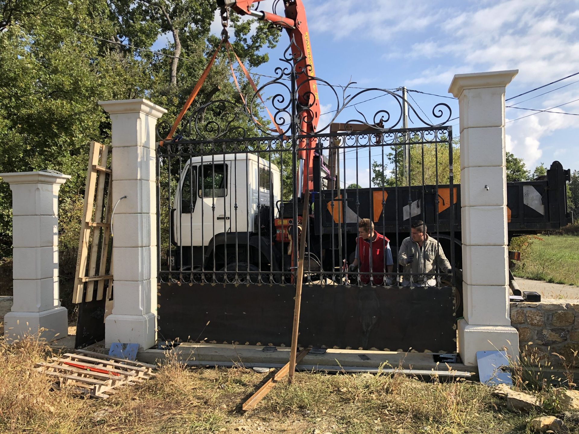 Un portail en fer forgé avec des piliers blancs, une grue et un camion derrière, et deux ouvriers à l'entrée.