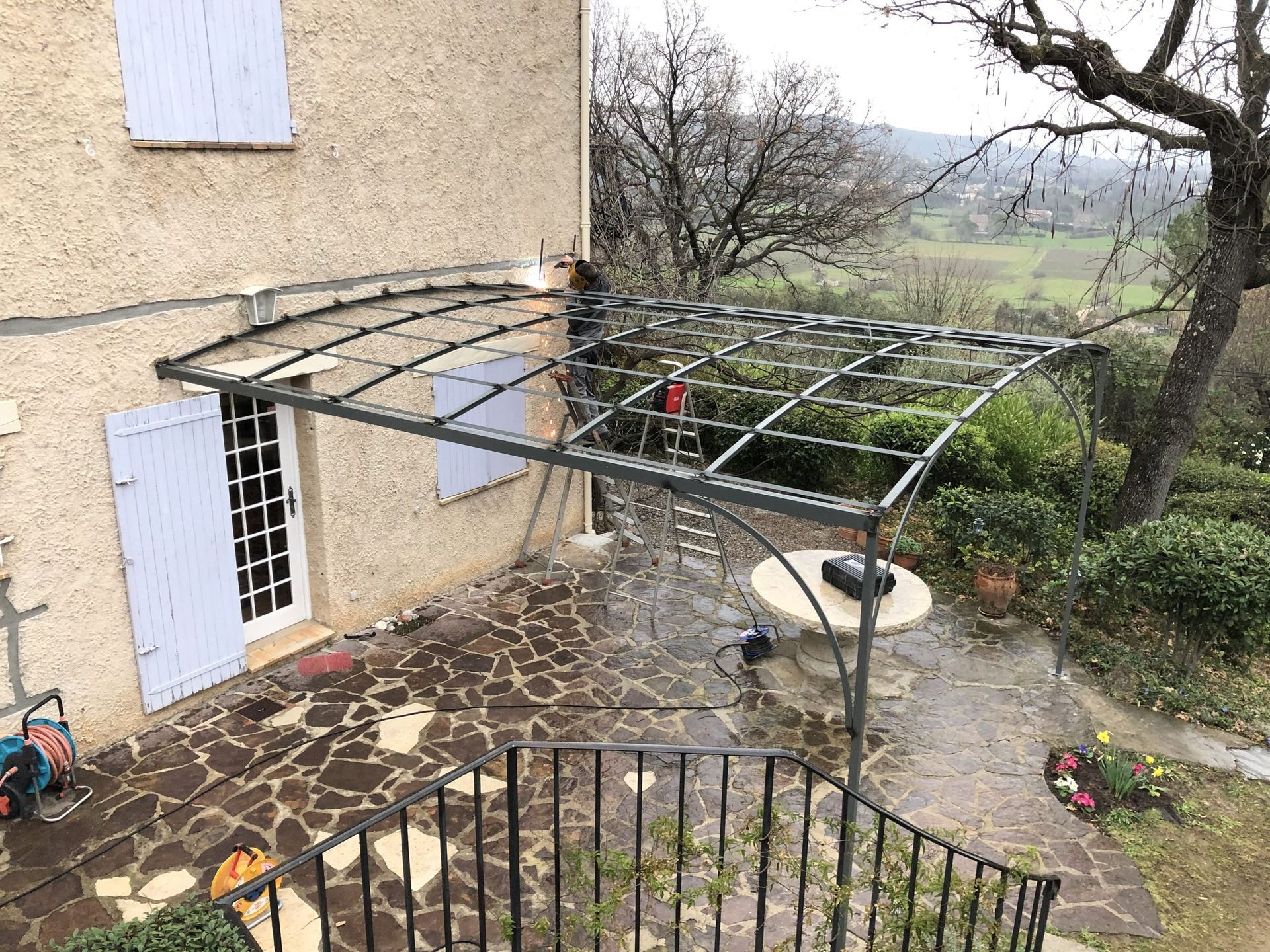 Terrasse arrière avec une pergola en métal à côté d'une maison, terrain humide et vue sur le jardin