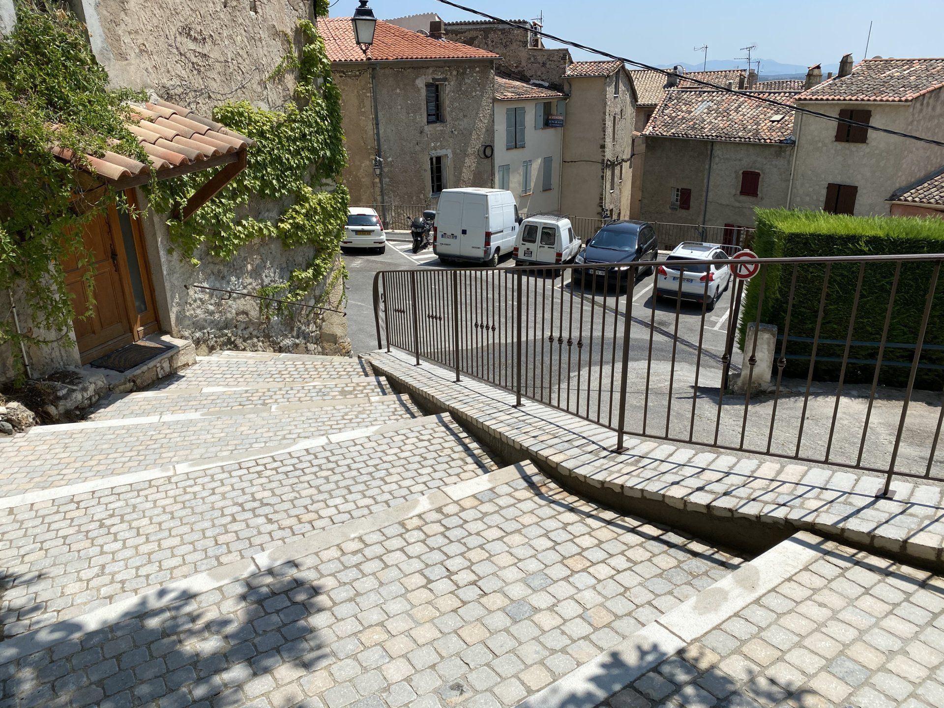 Ruelle pavée bordée de bâtiments en pierre, avec des voitures garées et une rambarde métallique baignée de soleil.