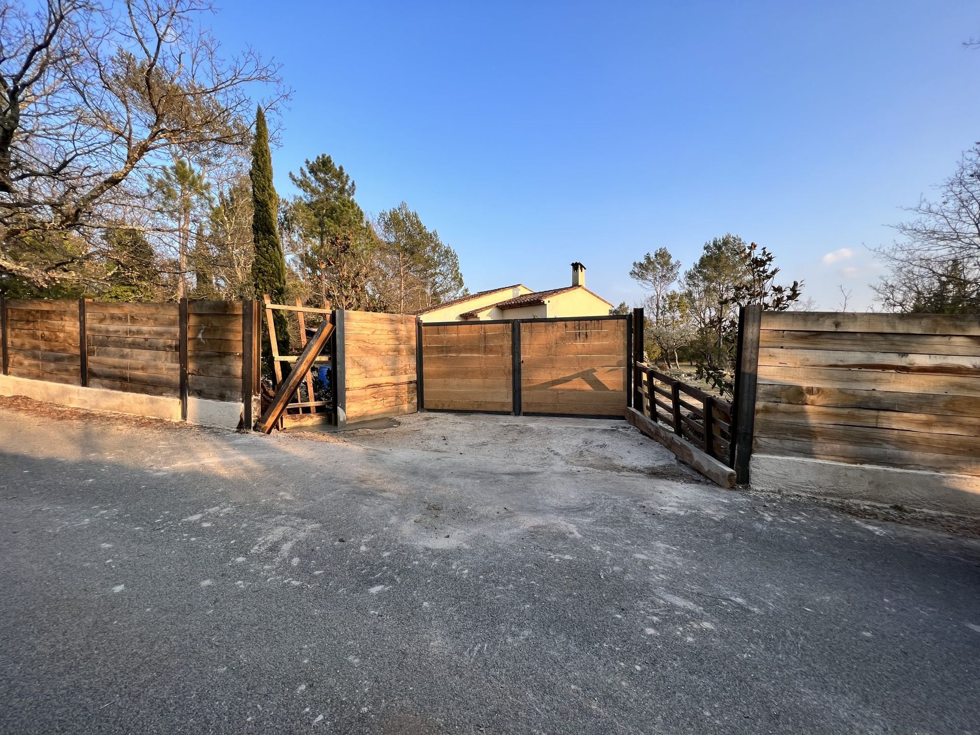 Allée de gravier avec portail en bois et clôture devant une maison sous un ciel bleu azur.