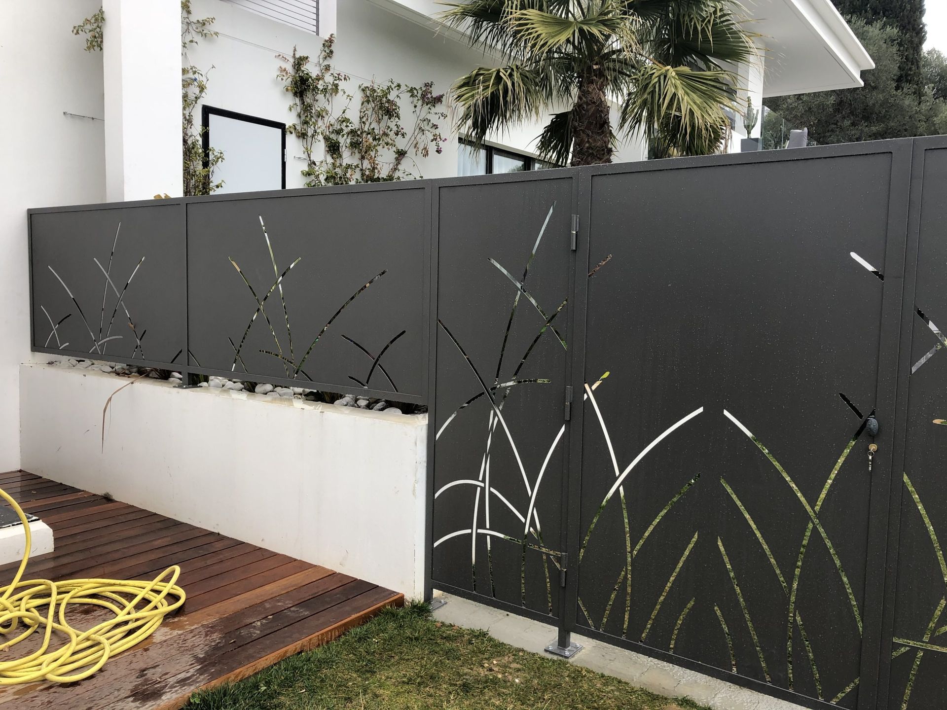 Clôture décorative gris foncé avec des découpes de plantes devant une maison blanche et un porche.