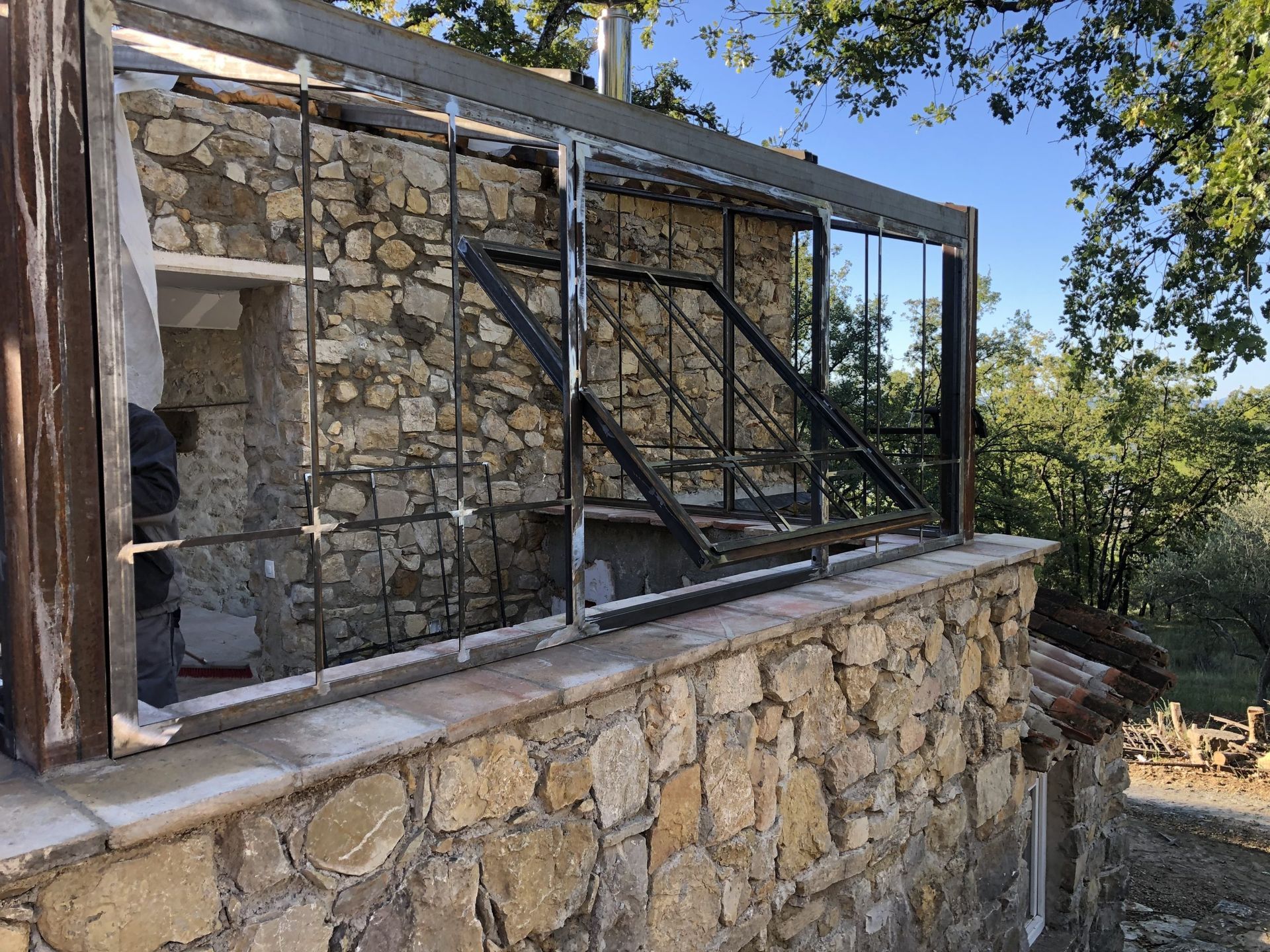 Bâtiment en pierre avec une grande fenêtre encadrée de métal, vu depuis une terrasse extérieure ensoleillée bordée d'arbres.