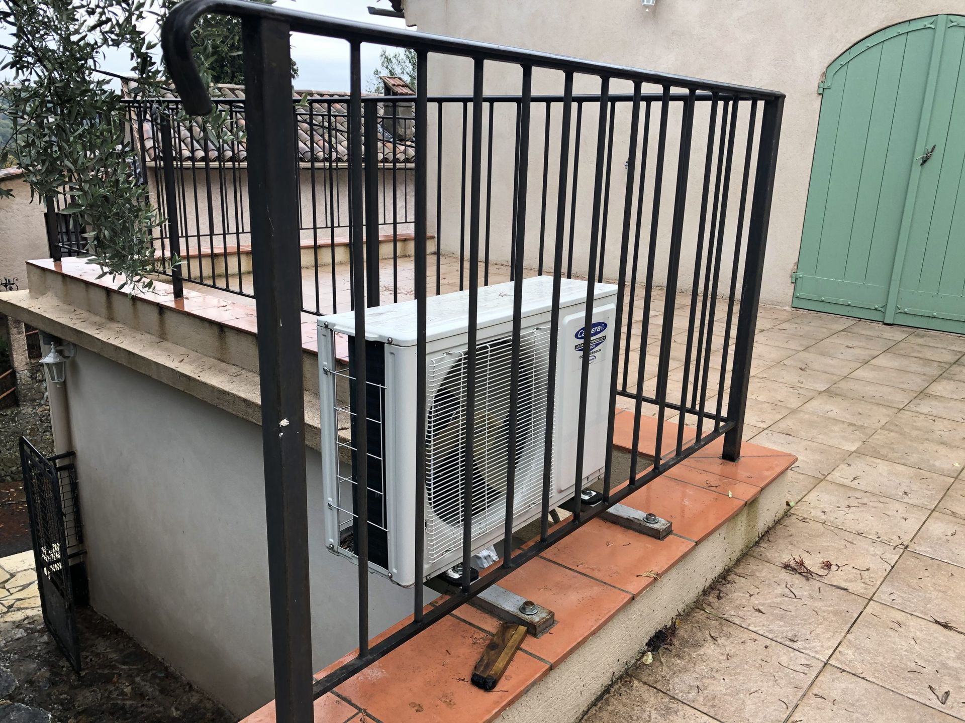 Unité de climatisation sur le toit, derrière une rambarde de sécurité en métal noir, sur une terrasse carrelée.