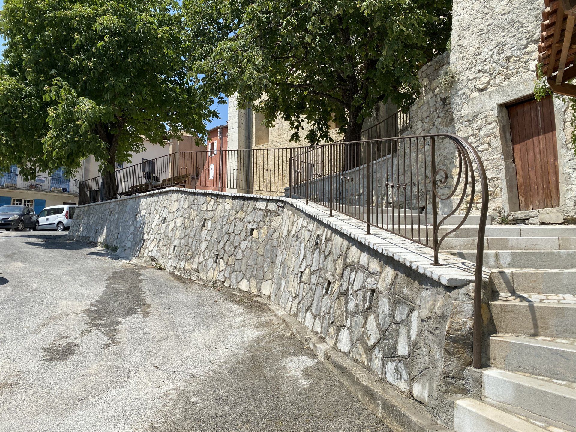 Mur en pierre et escaliers le long d'une rue ensoleillée bordée d'arbres et de bâtiments