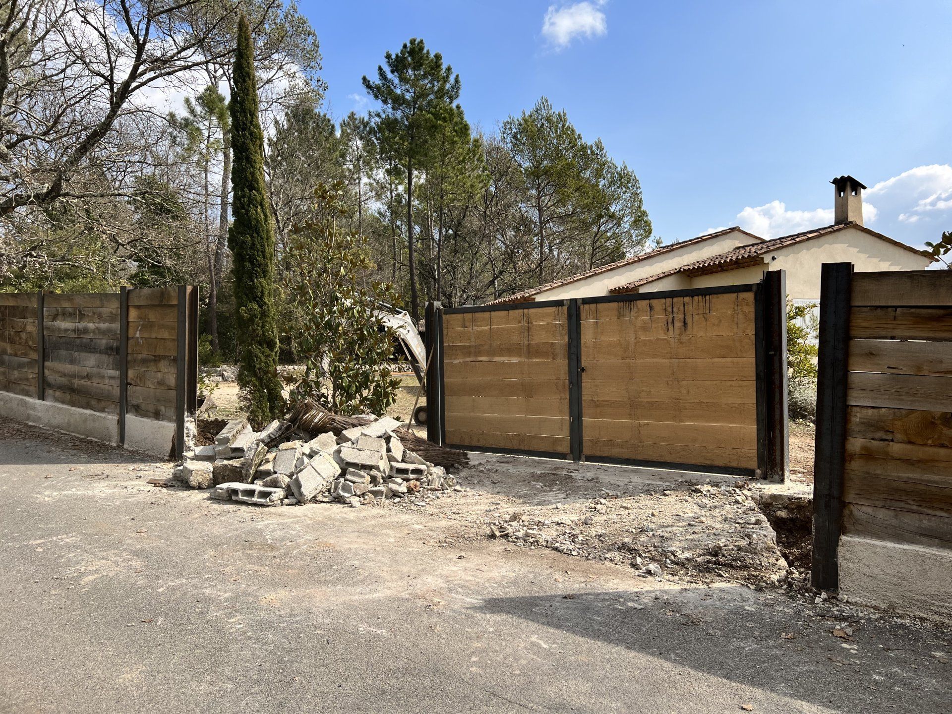 Clôture et portail en bois donnant sur une allée de campagne, près d'une maison et d'arbres, par une journée ensoleillée.