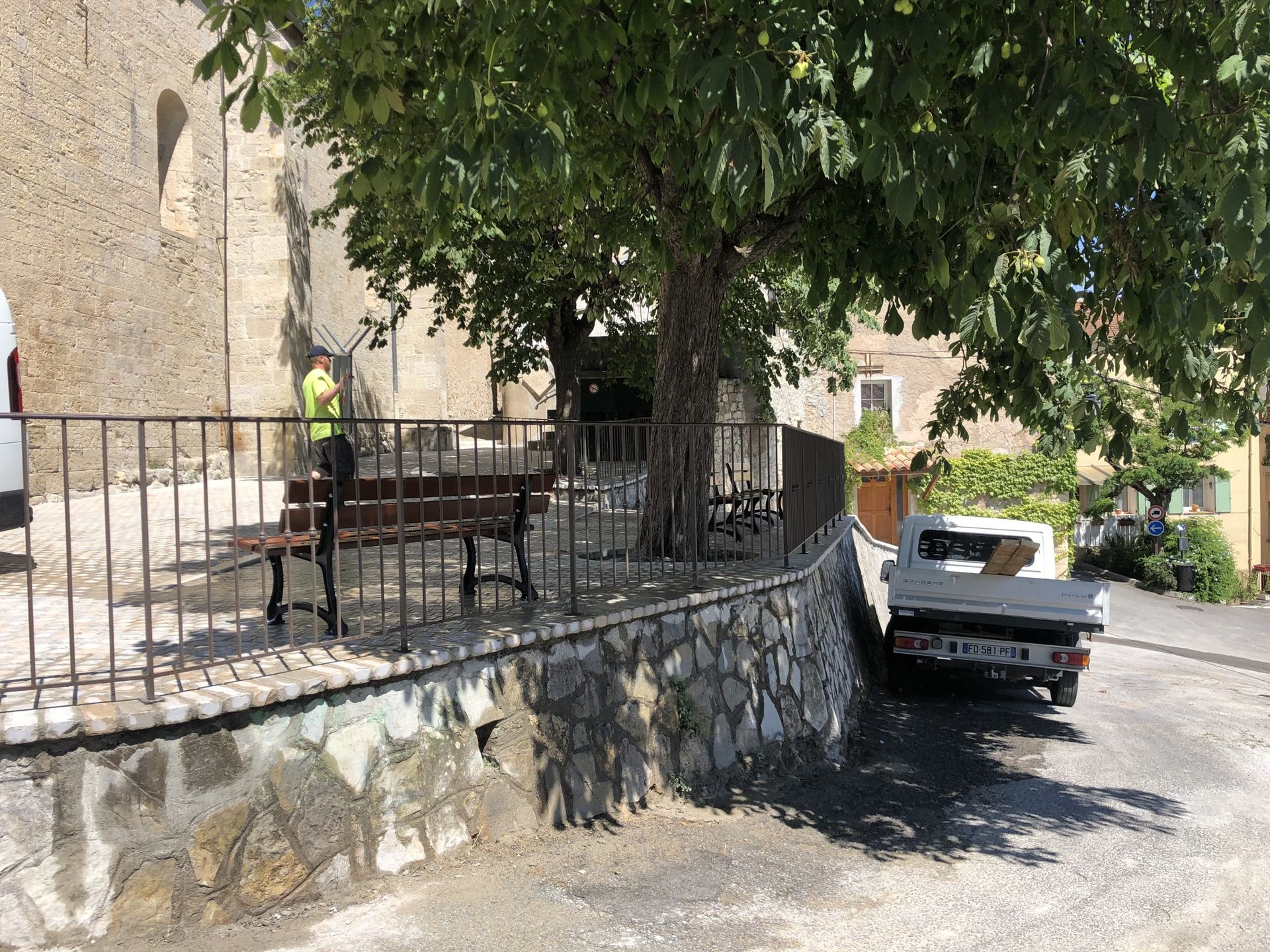 Terrasse en pierre avec rambarde noire sous un arbre, une camionnette blanche garée à côté en plein soleil.