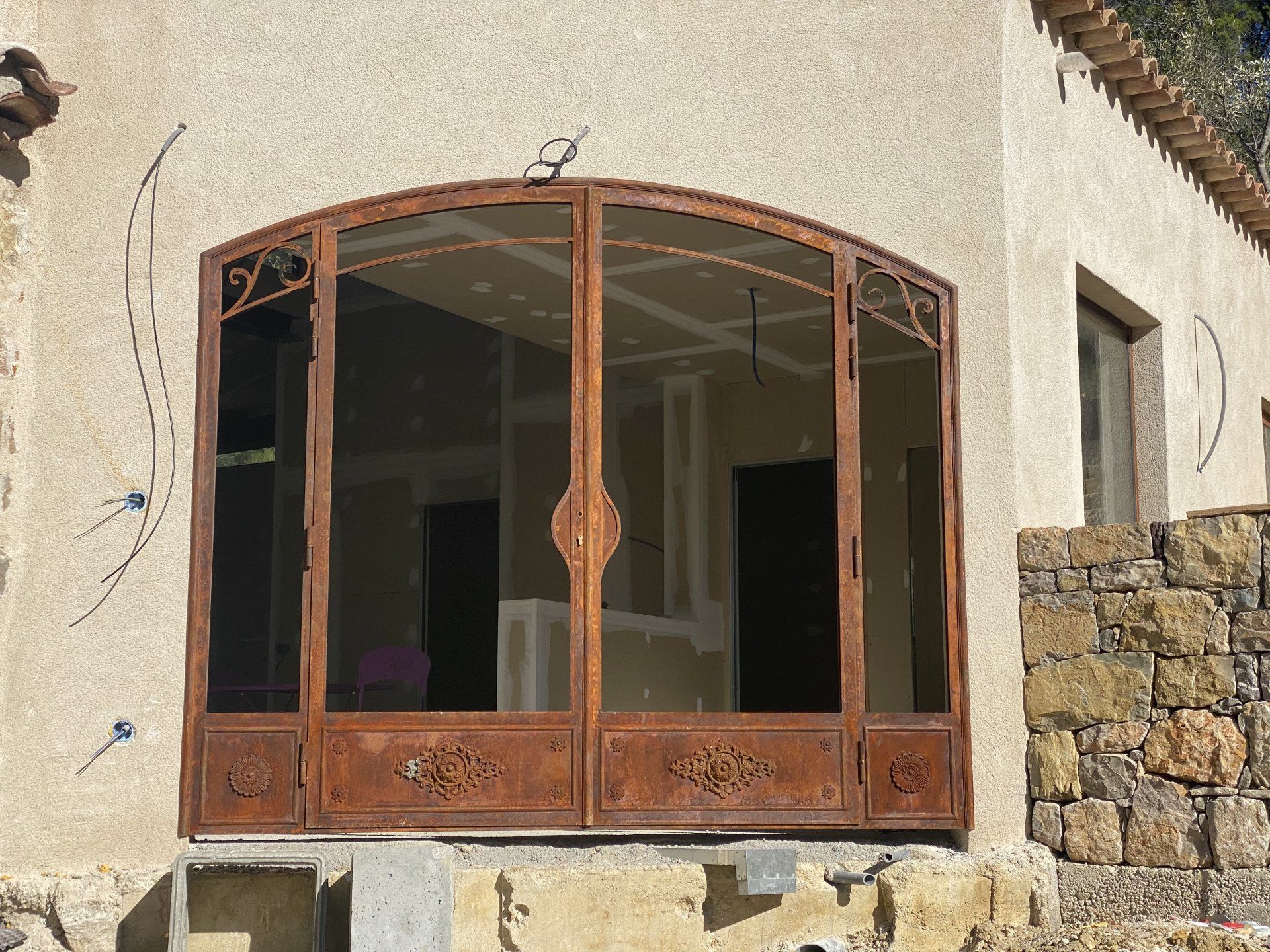Fenêtre cintrée couleur rouille dans un mur extérieur en pierre et en stuc, révélant une pièce inachevée à l'intérieur.