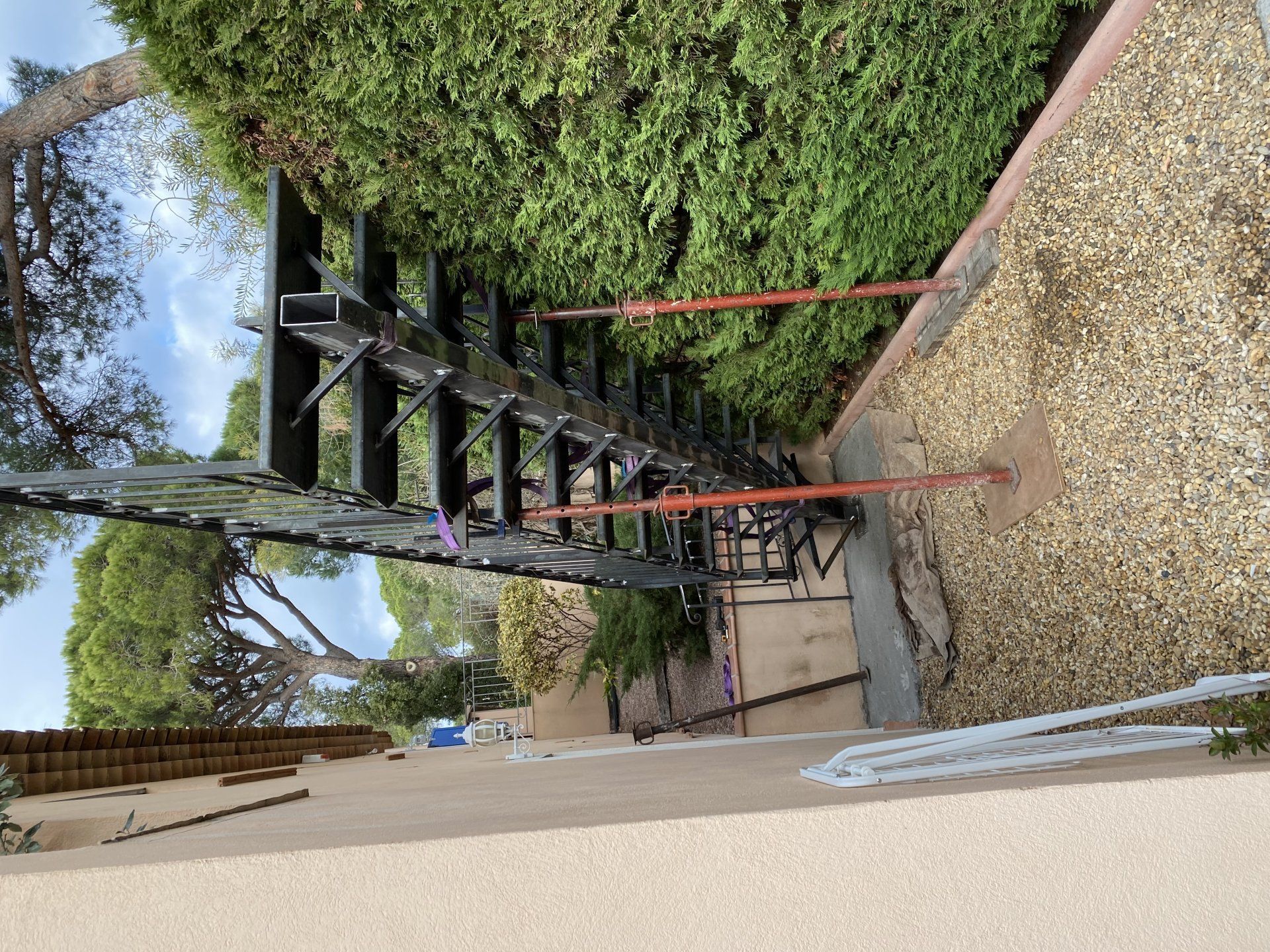 Petit escalier en métal noir au bord d'un chemin de gravier, avec des rampes rouges et des arbres feuillus au-dessus.