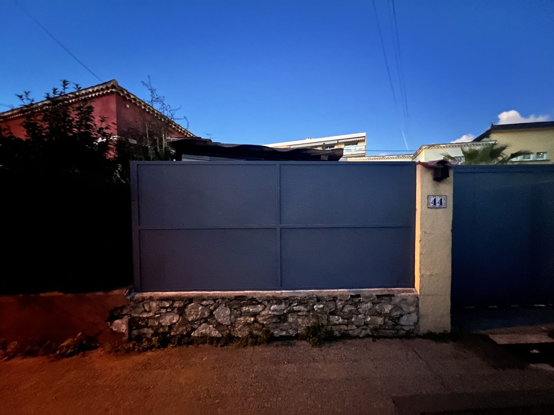 Portail en métal bleu et mur en pierre devant une maison dans une rue calme.