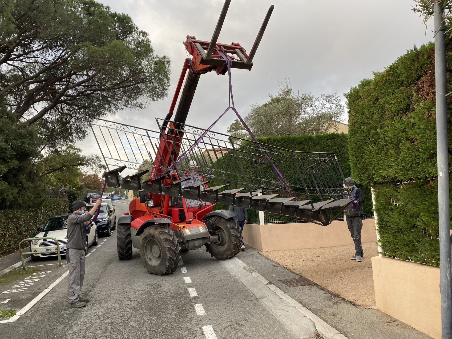 Une grue rouge soulève un grand panneau de clôture noire au-dessus d'une route, des ouvriers se trouvant à proximité, dans un quartier résidentiel.