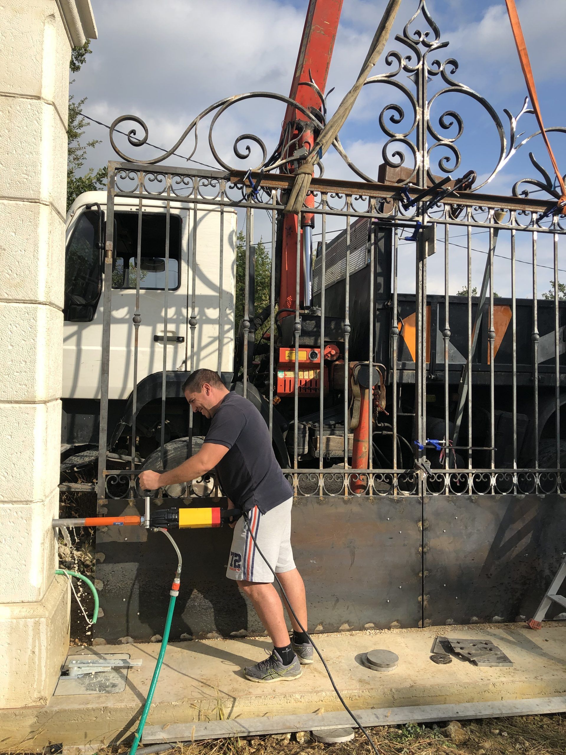 Un ouvrier utilise une scie électrique à côté d'un portail en fer forgé orné, une grue étant visible derrière lui.