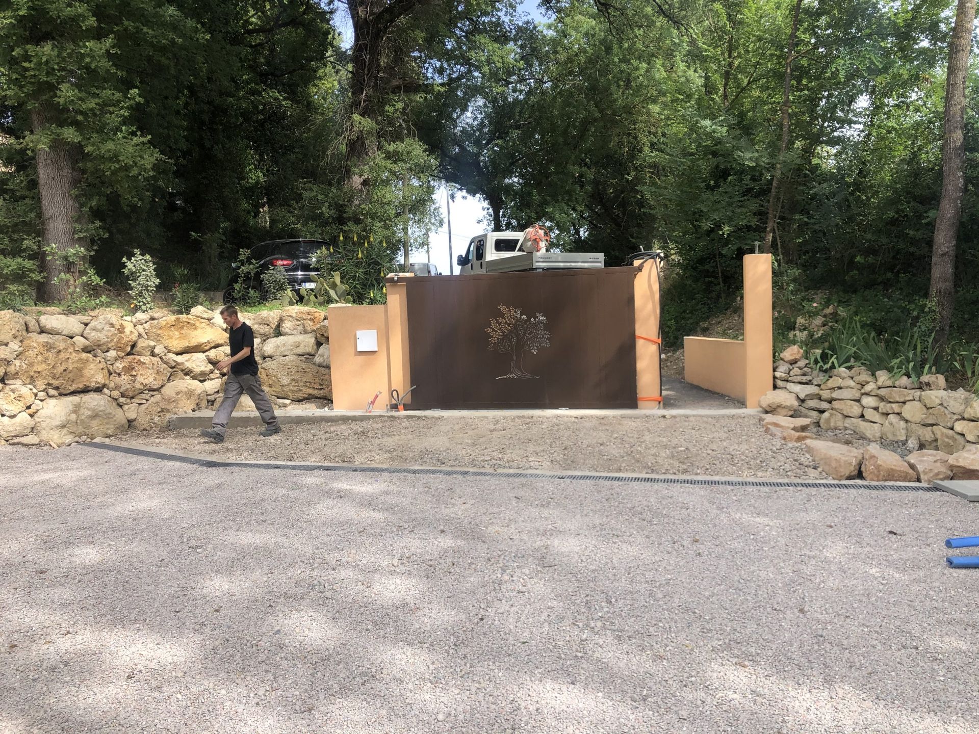 Une personne passe devant un portail marron, entre des murs de pierre et des arbres, au bord d'un chemin de gravier.