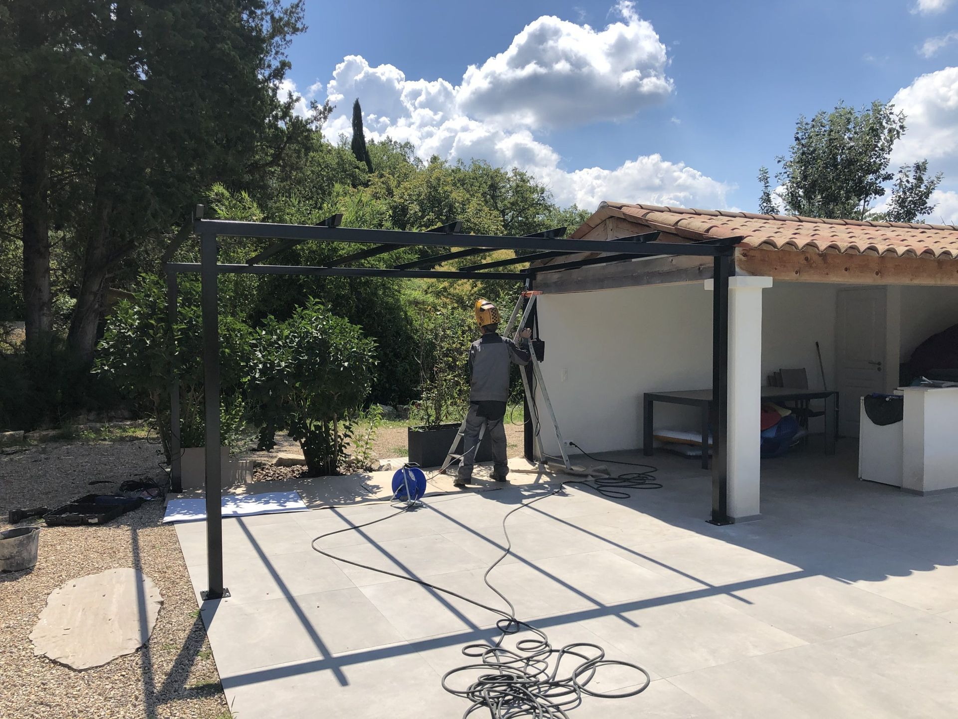 Une personne installe une structure de pergola en métal noir sur une terrasse ensoleillée à côté d'un garage blanc.