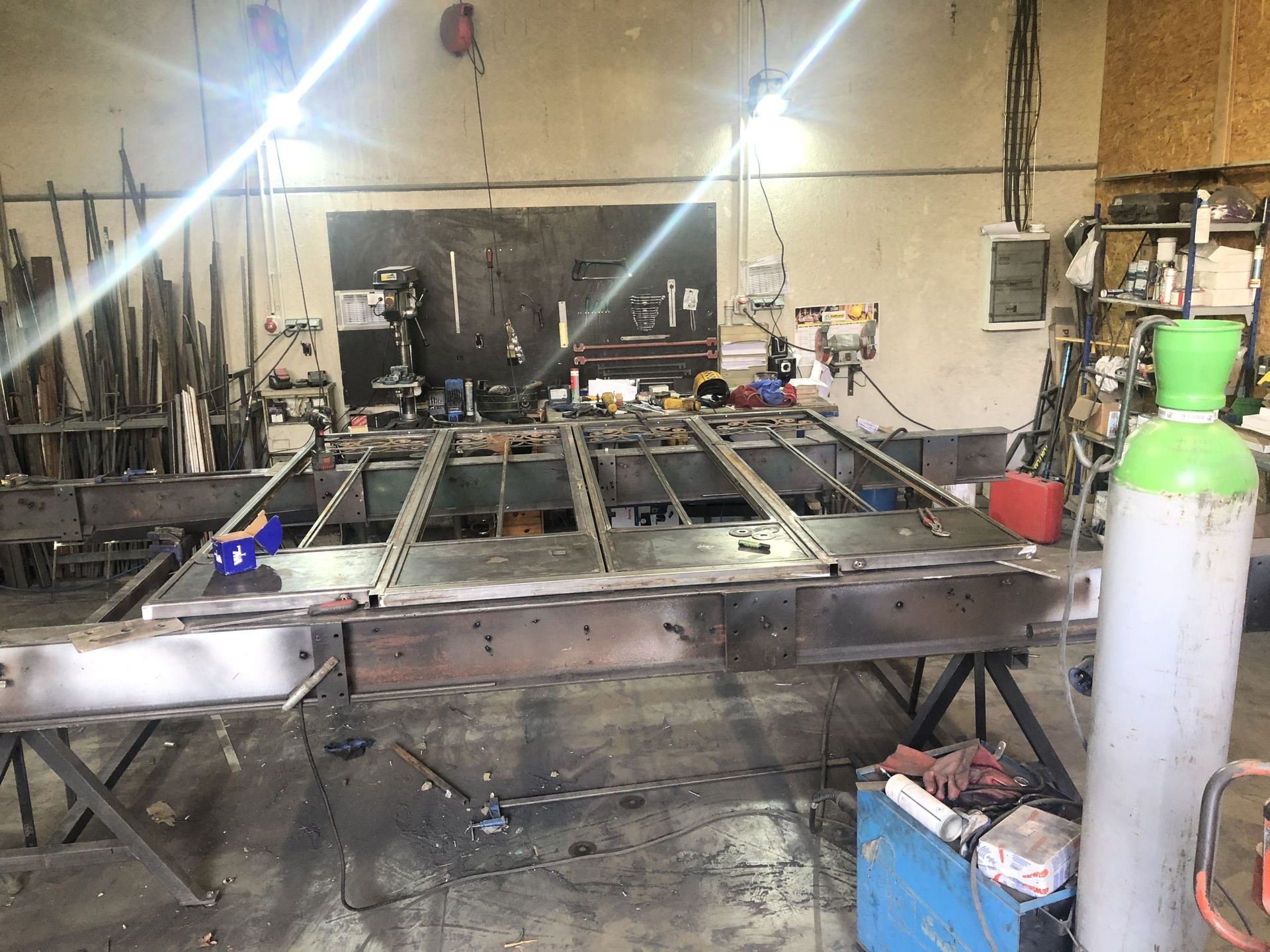 Atelier sombre avec une grande table de fabrication métallique, des outils et une bonbonne de gaz verte au premier plan.