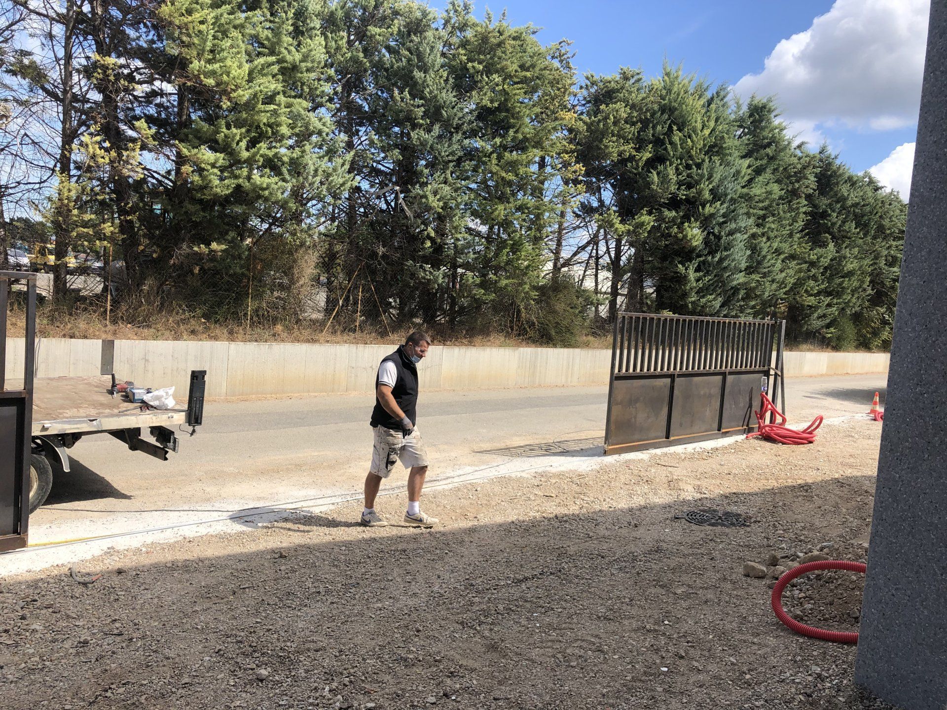 Une personne traverse un terrain gravillonné le long d'un mur en béton et d'arbres, avec du matériel rouge sur sa droite.