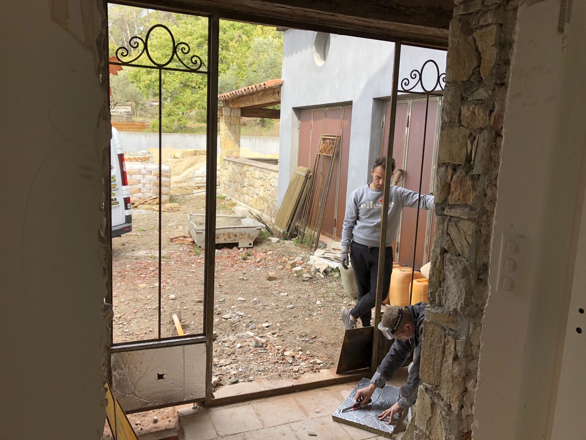Deux personnes travaillent près d'une porte dans une cour partiellement démolie, jonchée de débris et aux murs apparents.