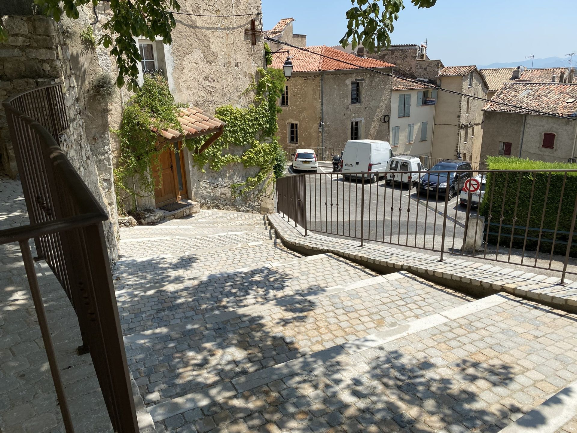 Rue pavée bordée de maisons en pierre et d'une petite place avec des voitures garées et l'ombre tachetée des arbres