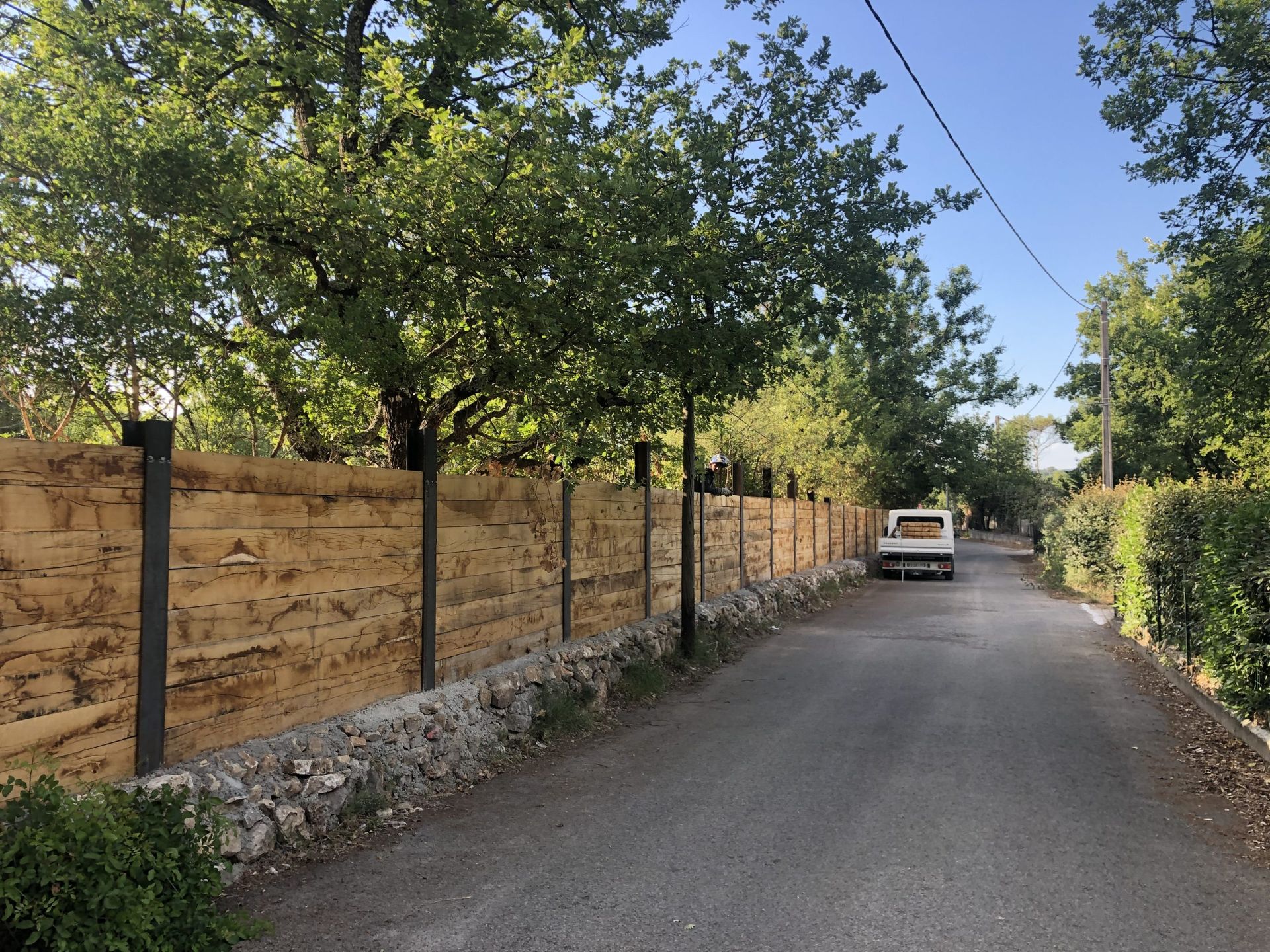 Allée de gravier bordée d'arbres, longeant un mur de pierres beiges, menant à un portail sous un ciel bleu.