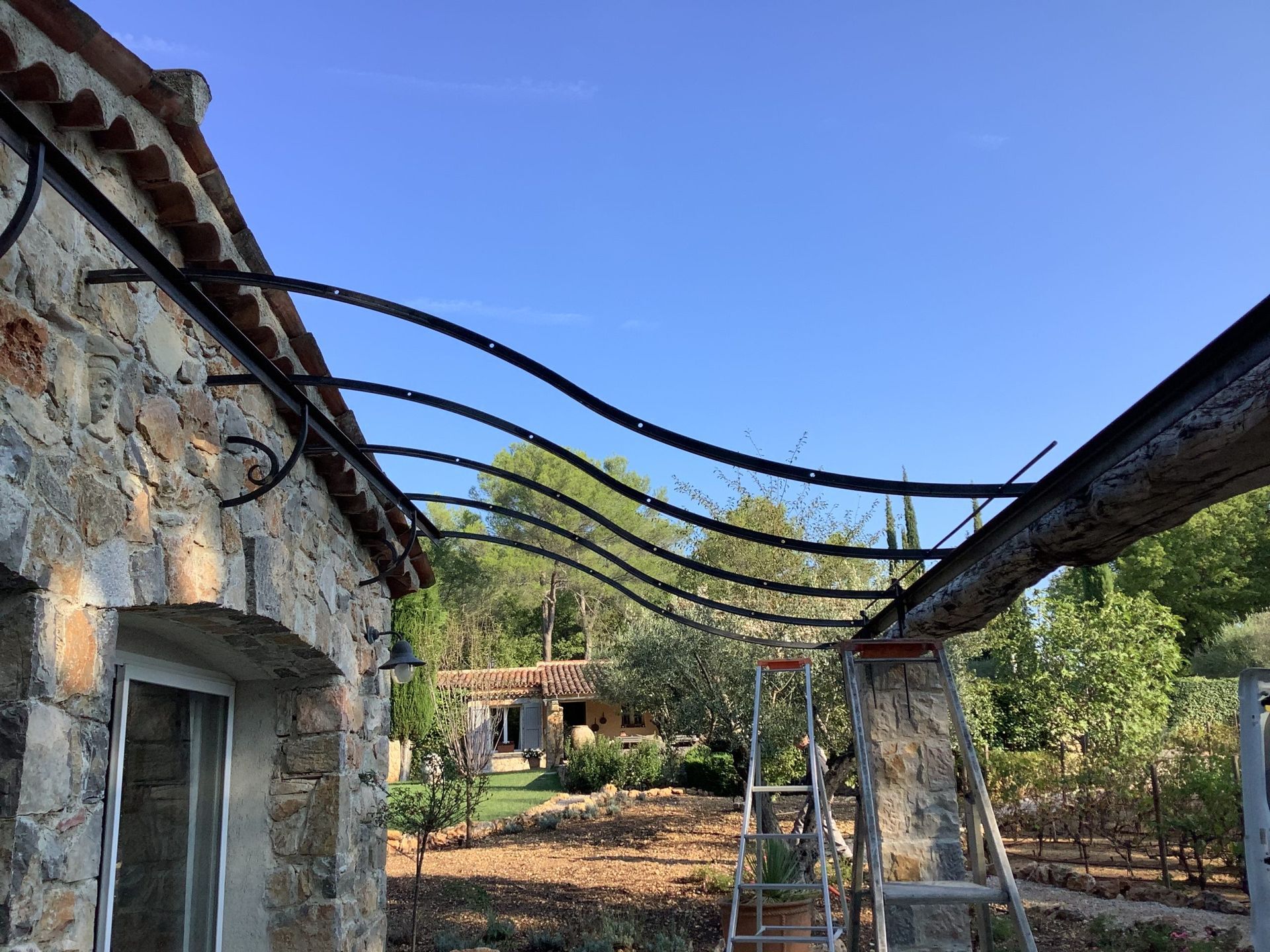 Bâtiments rustiques en pierre avec une passerelle en treillis métallique incurvée sous un ciel bleu, entourés d'arbres.