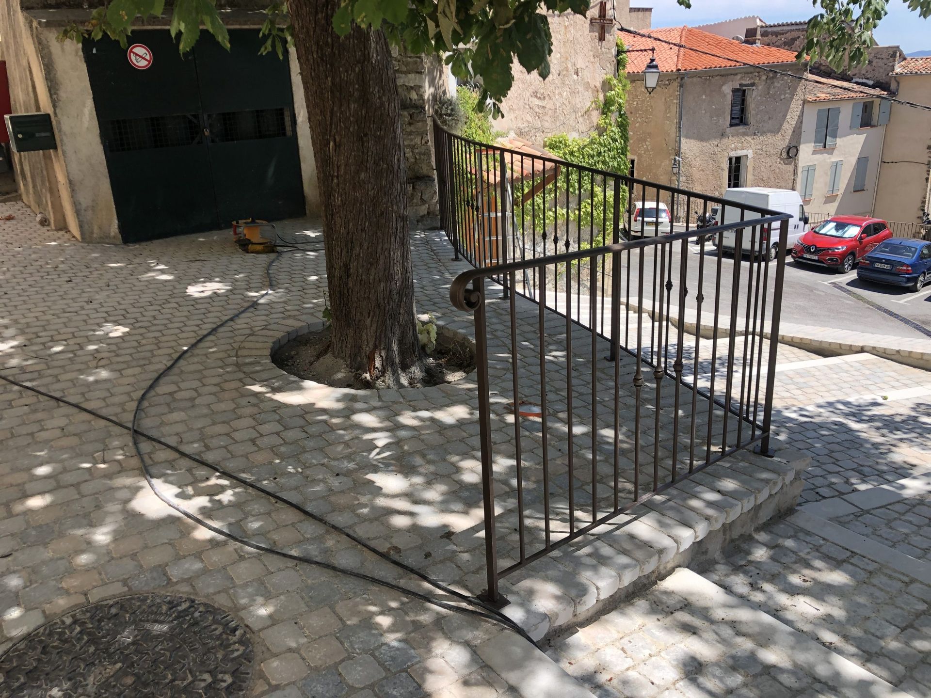 Allée en pierre avec un arbre et une rambarde en métal noir, longeant une rue en pente et des bâtiments en pierre.