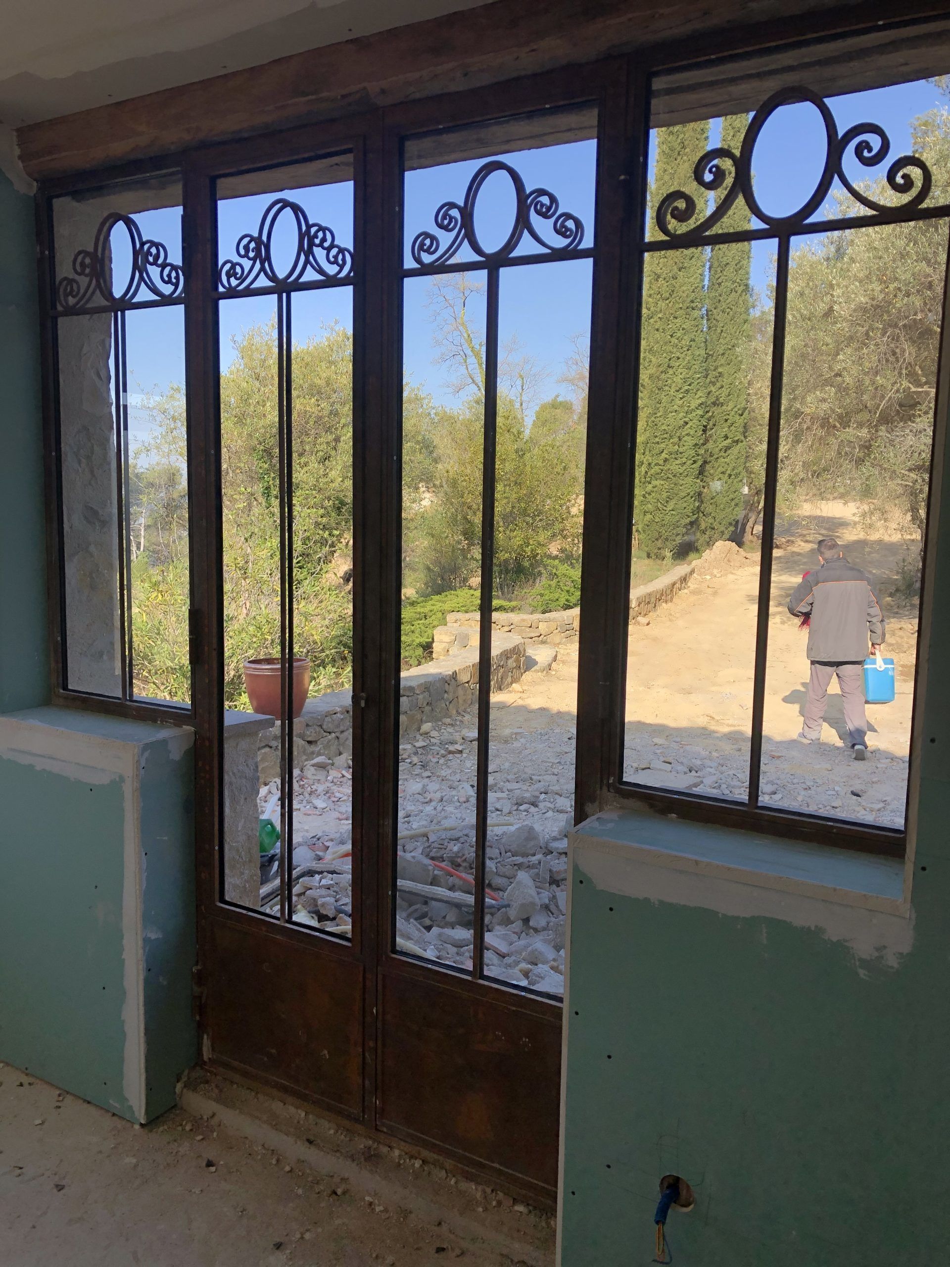 Vue depuis le porche, à travers la porte vitrée et les fenêtres, sur une cour boisée et ensoleillée, avec une personne qui marche à l'extérieur.
