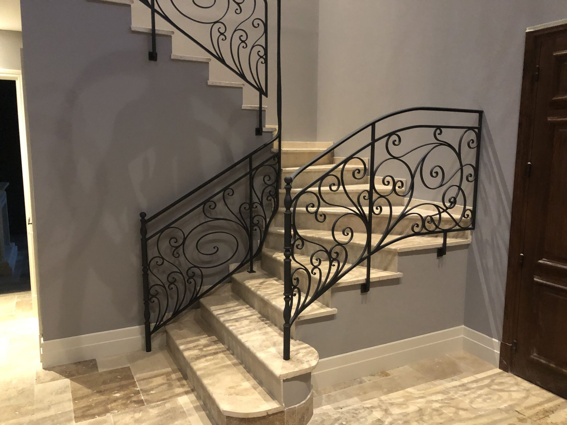 Élégant escalier intérieur avec rampes en fer forgé noir et marches en marbre crème, jouxtant une porte en bois.