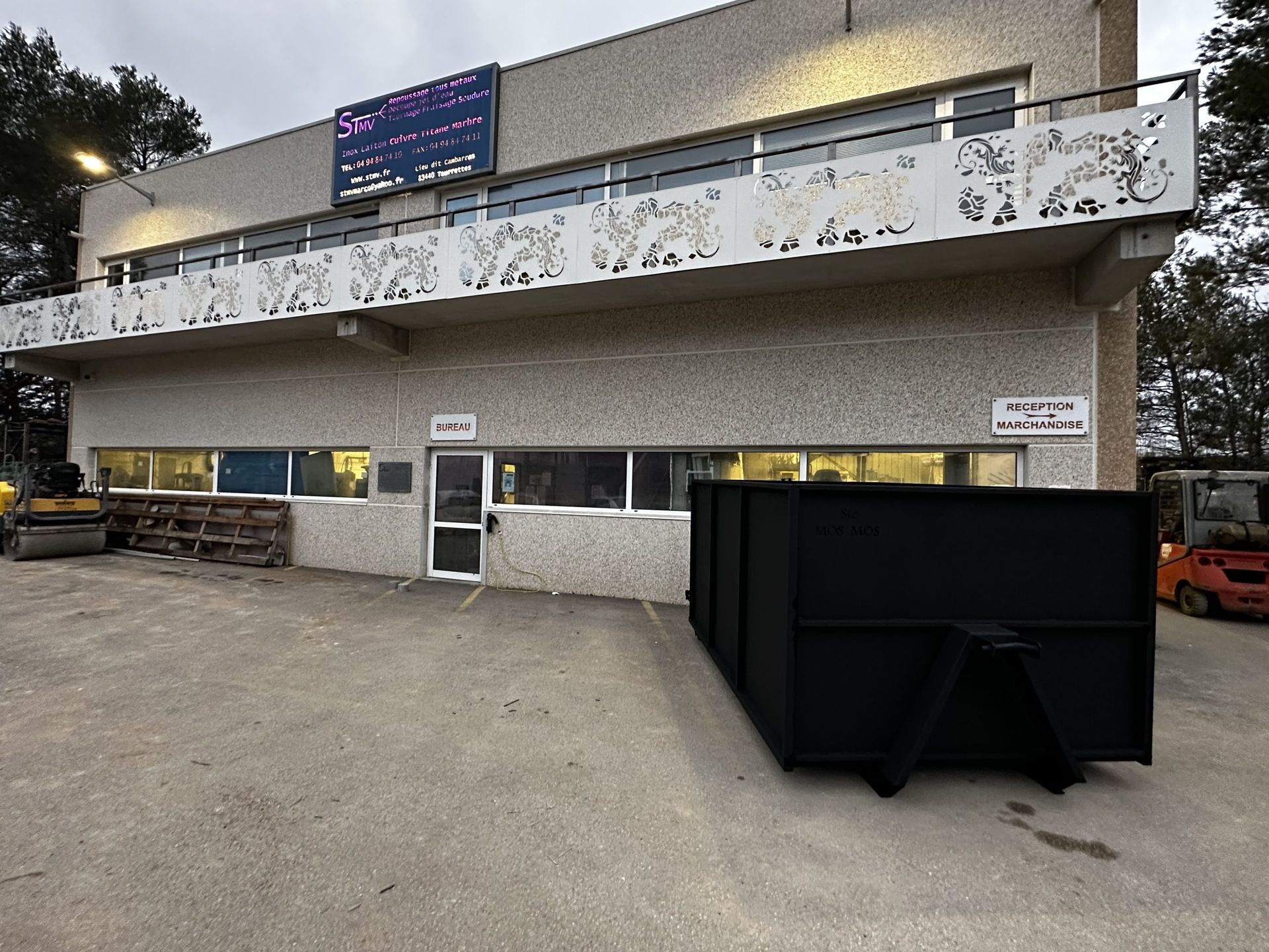 Extérieur d'un bâtiment de deux étages avec balustrade de balcon blanche, fenêtres éclairées et engins de chantier stationnés à l'extérieur